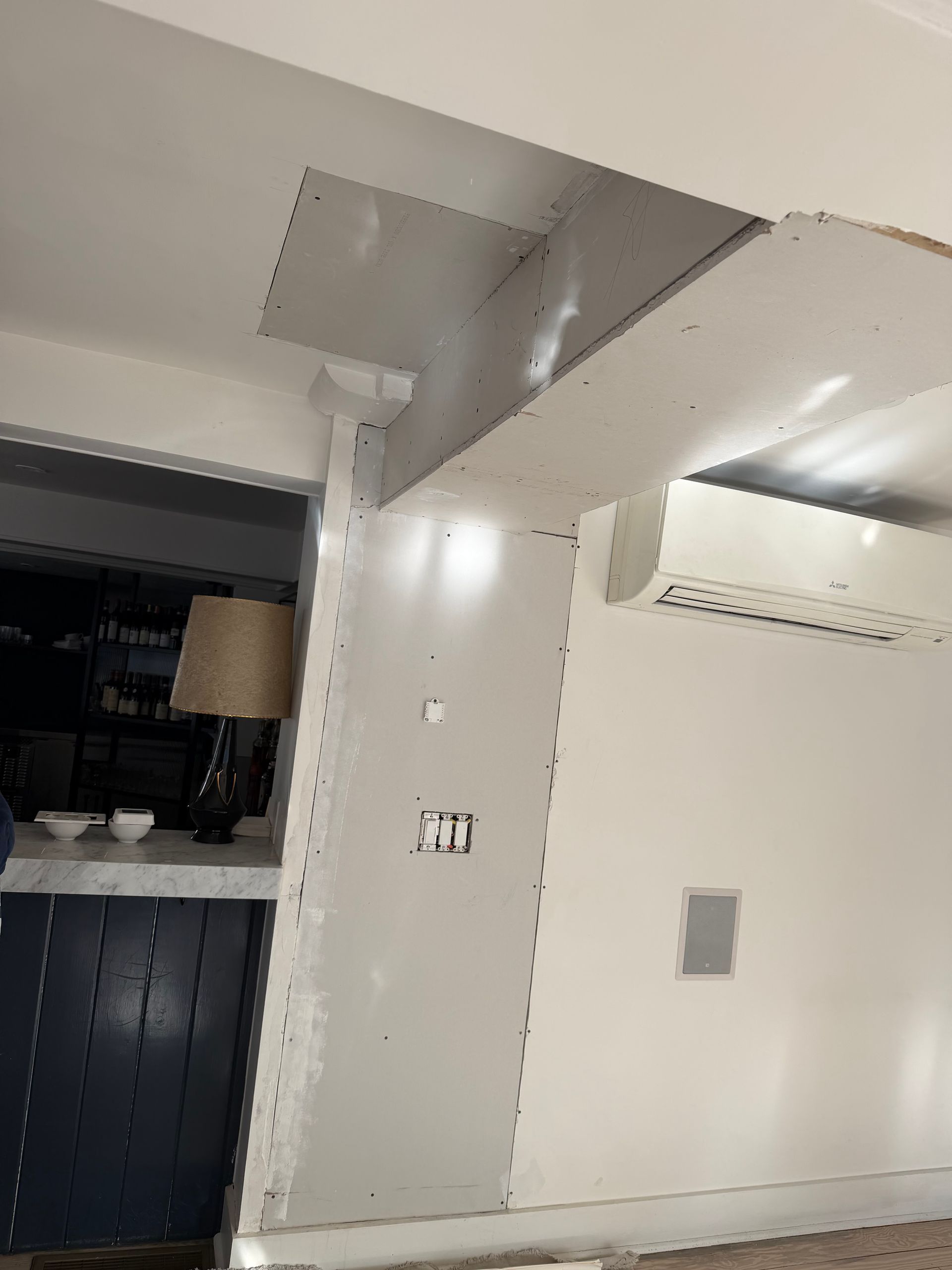 A partially finished drywall renovation on a wall and ceiling corner near an indoor air conditioning unit.