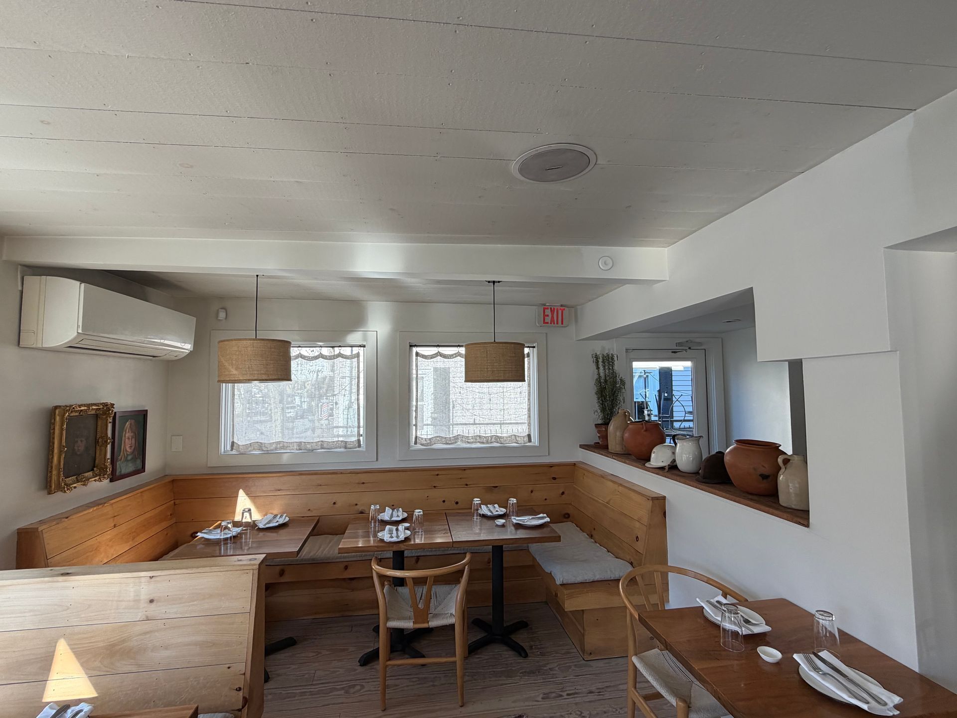 A dining room with wooden bench seating, tables, chairs, and lamps in a white-walled space with a window.