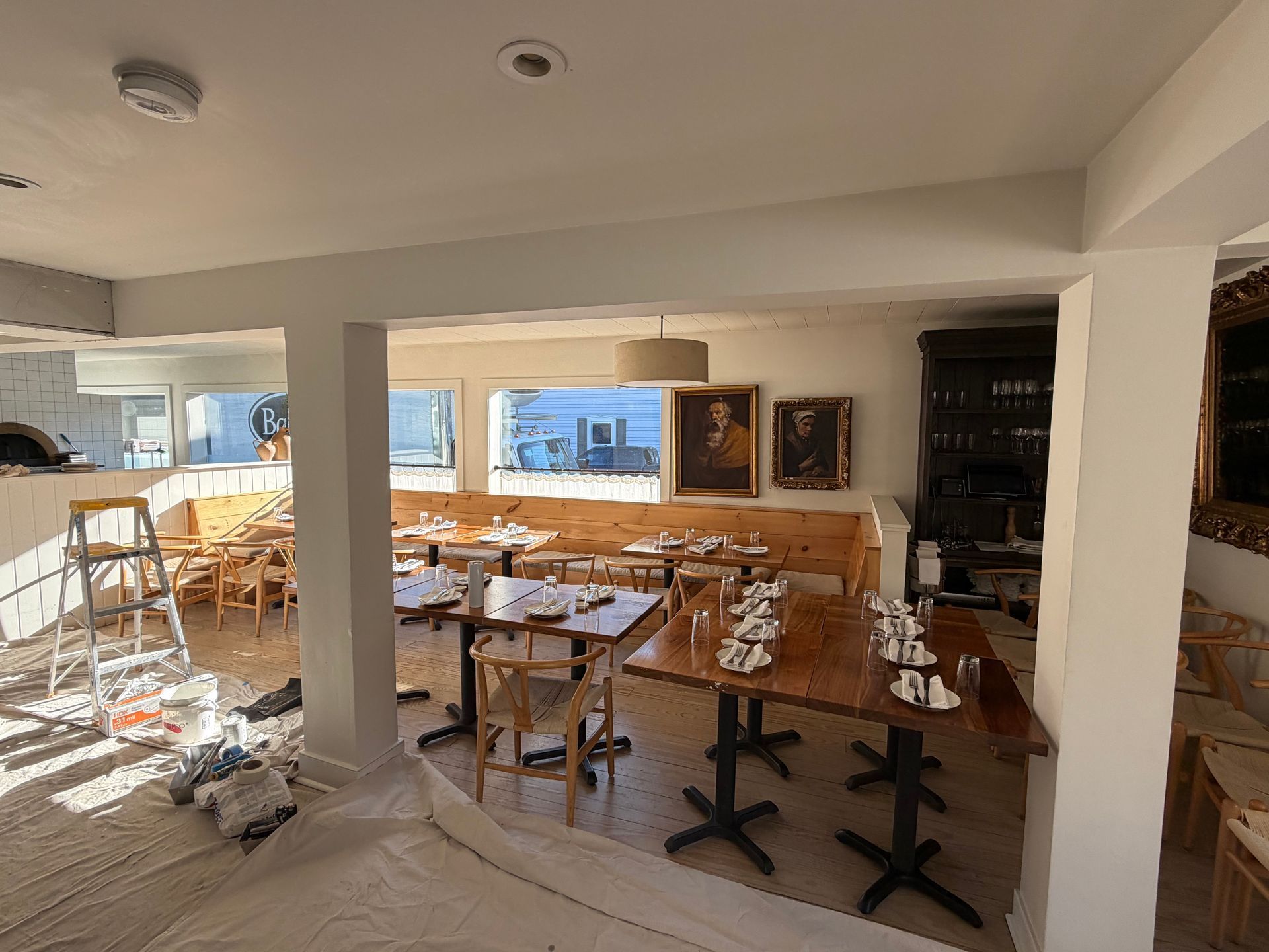 A dining area during renovation with drop cloths, a ladder, tables arranged in rows, and framed art on a white wall.