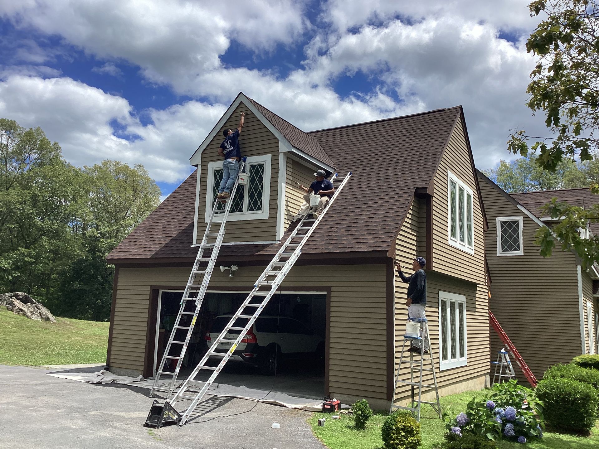 Three workers on ladders painting a brown house exterior; blue sky overhead.