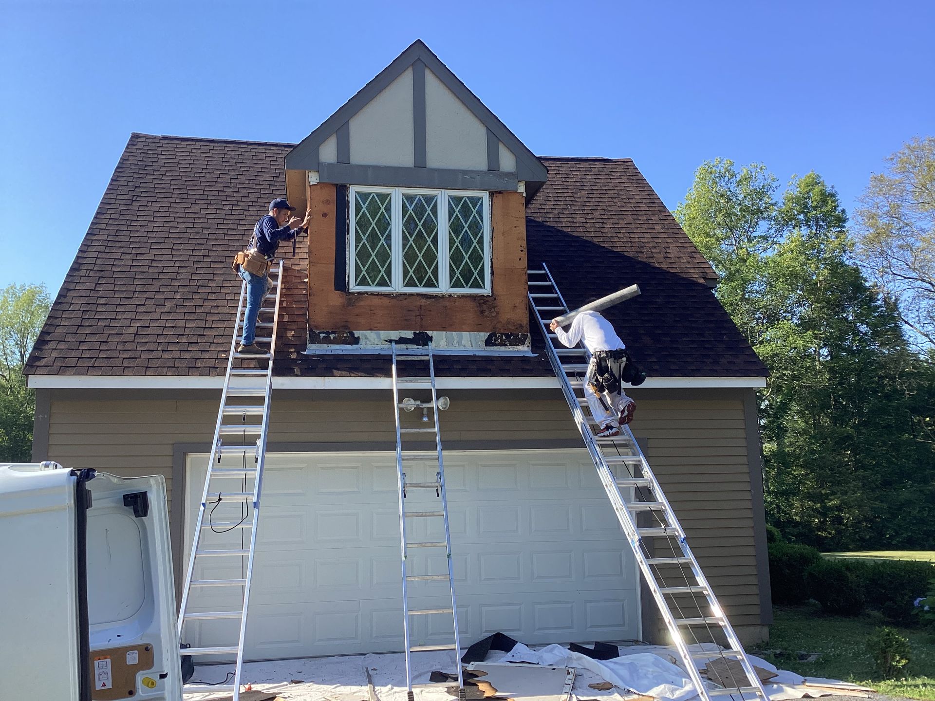 Two workers on ladders repair a garage exterior, near a window under a dormer.