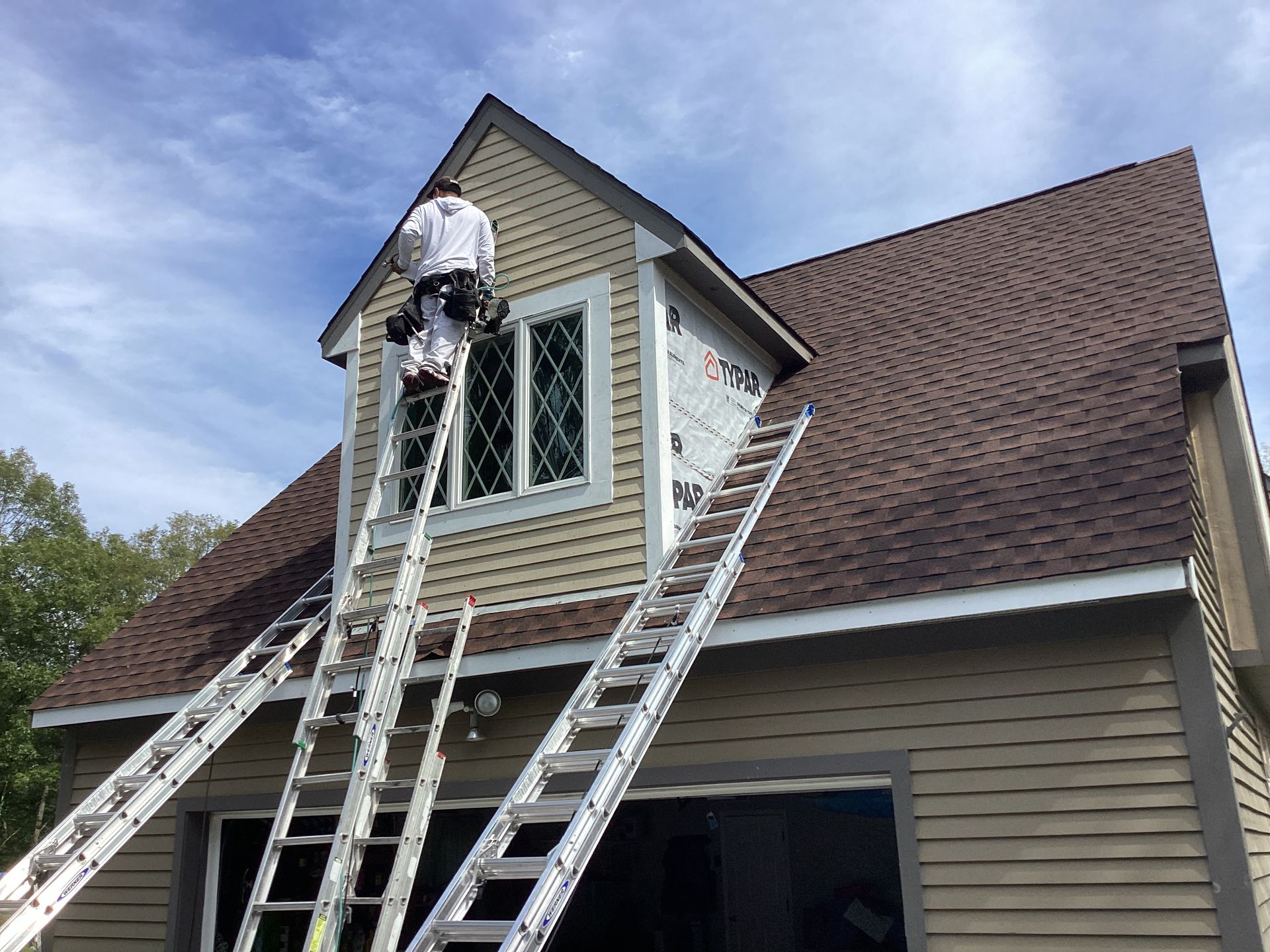 Person on a ladder painting a dormer window on a two-story tan house with brown roof. Blue sky in background.