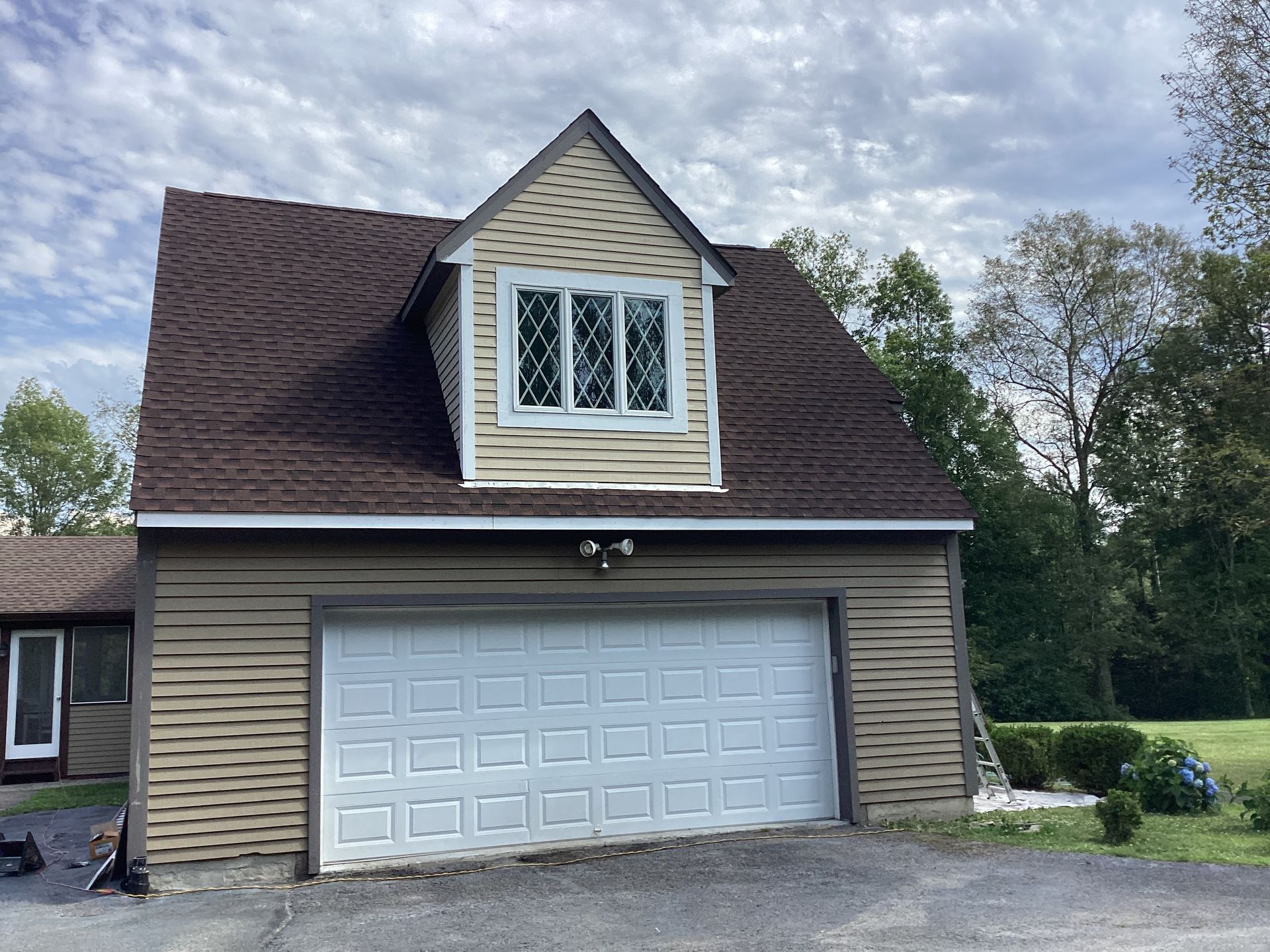 Tan garage with a white door, dormer with window, brown roof, and cloudy sky.