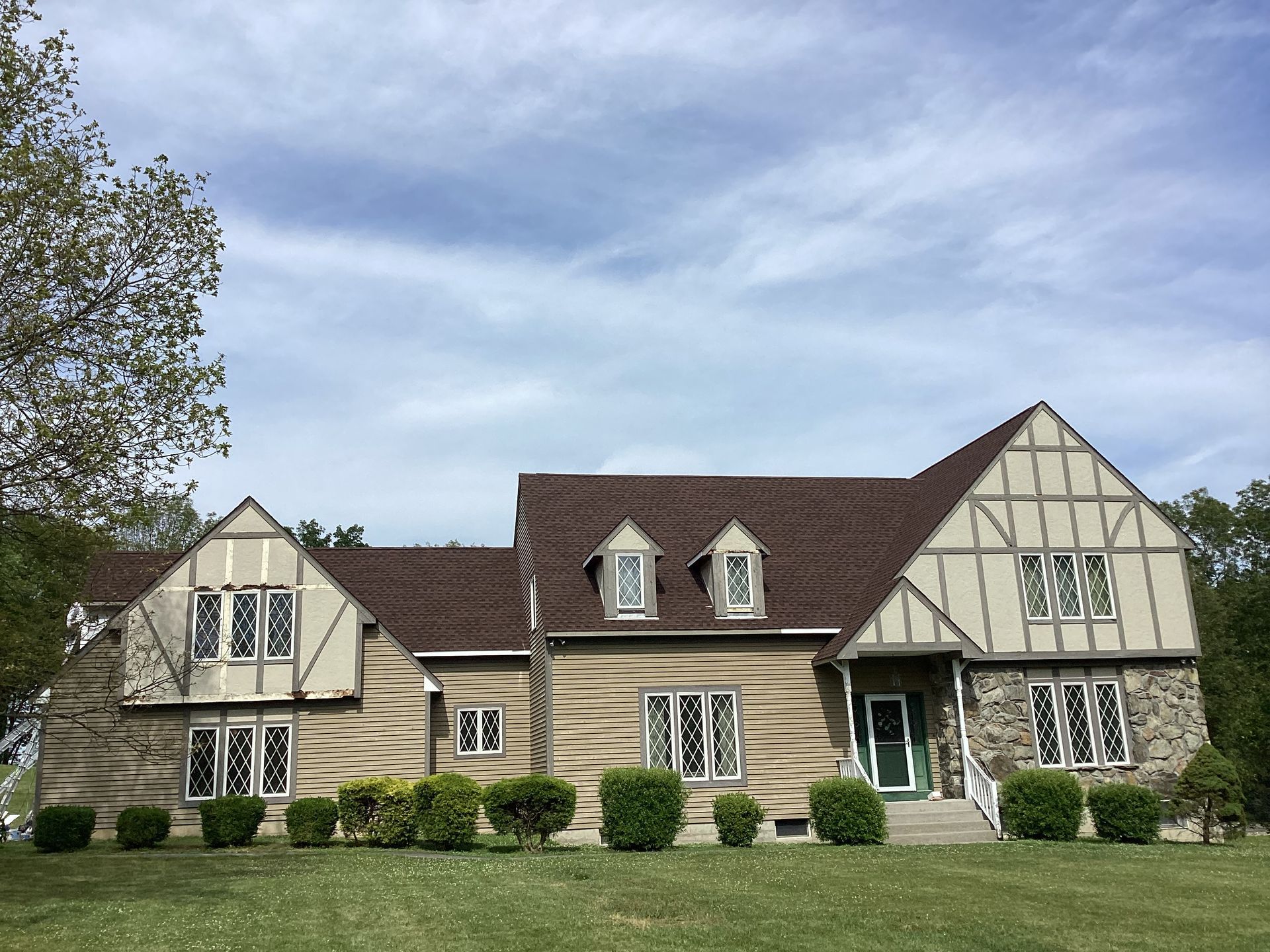 Tudor-style house with beige walls, brown roof, and manicured lawn under a partly cloudy sky.