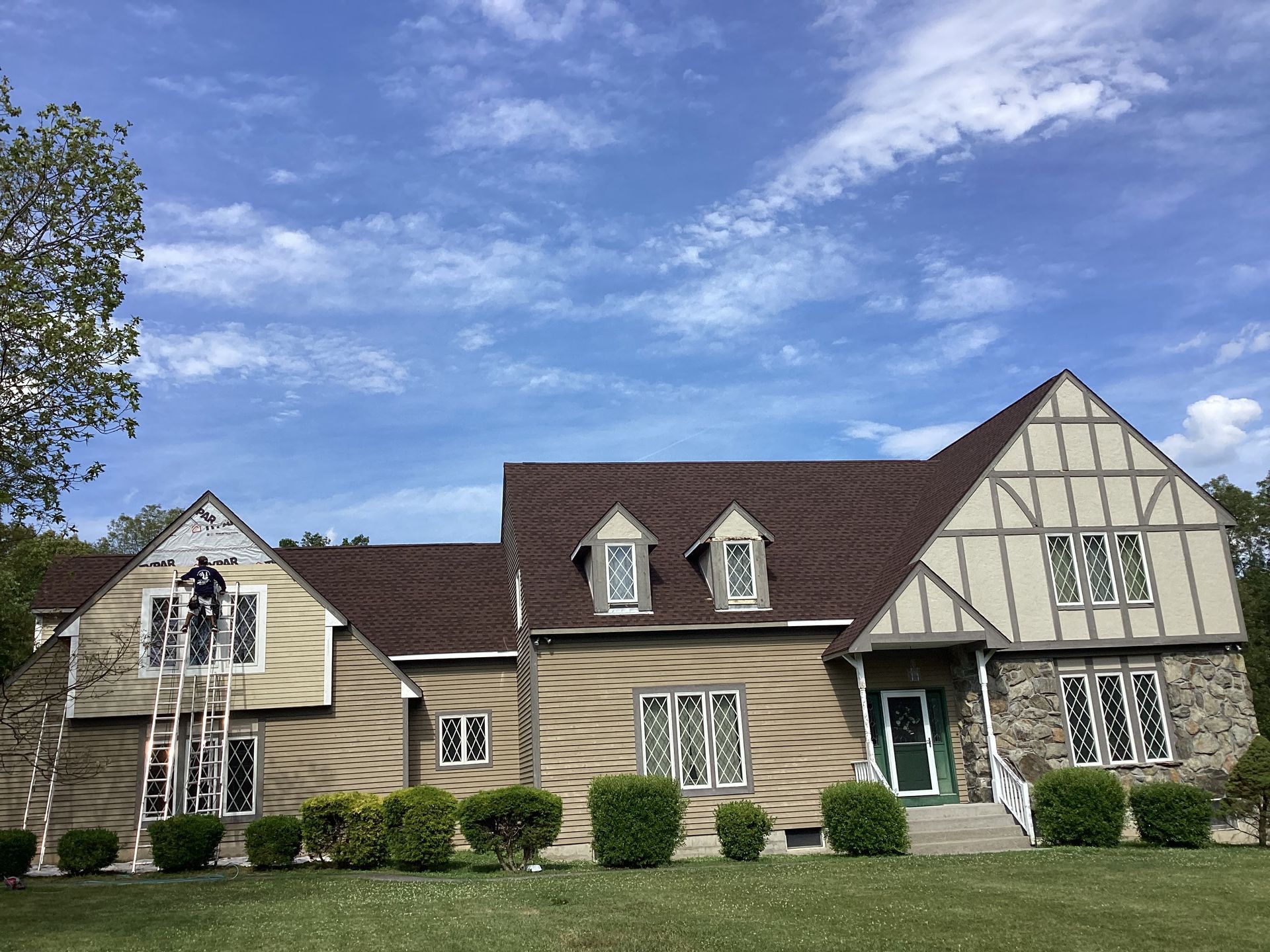 House with brown roof, beige walls, and Tudor-style trim. A worker on a ladder repairs the siding under a blue sky.
