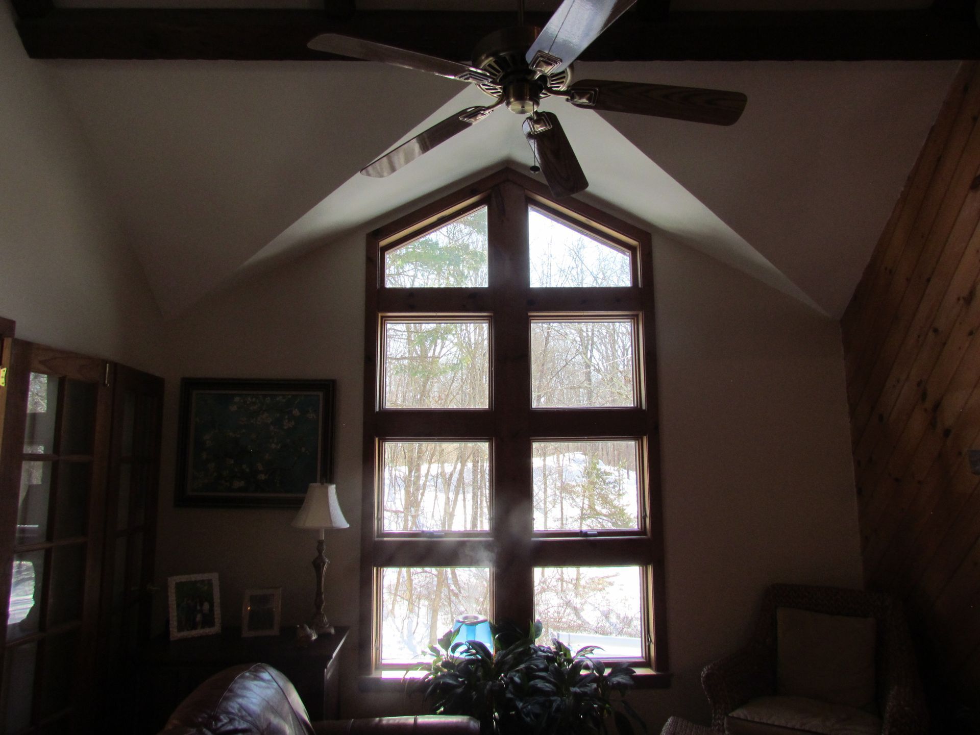 High-ceiling room with a large window, ceiling fan, and wooden accents. Sunlight streams through the window.