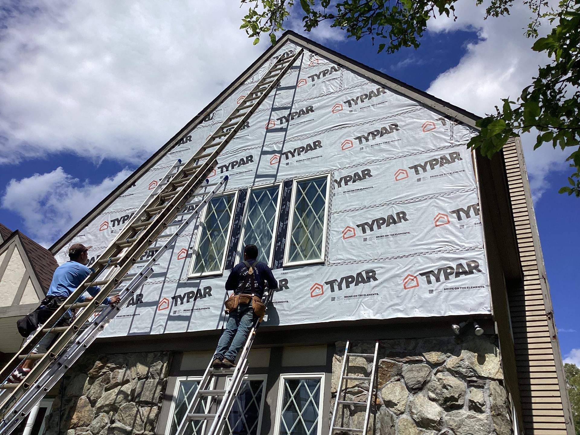 Two people installing Typar house wrap on a two-story house with stone facade and windows.