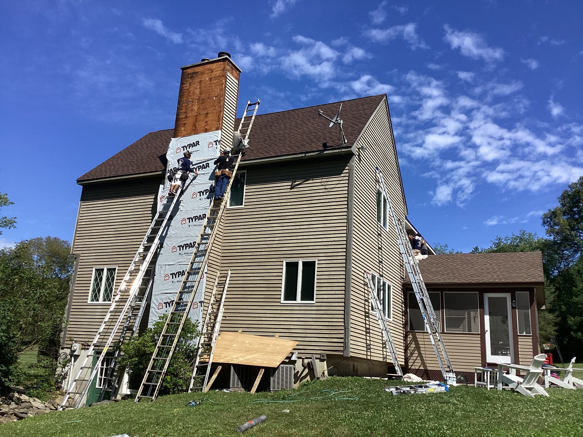 Workers on ladders repairing a house, removing siding near the chimney on a sunny day.