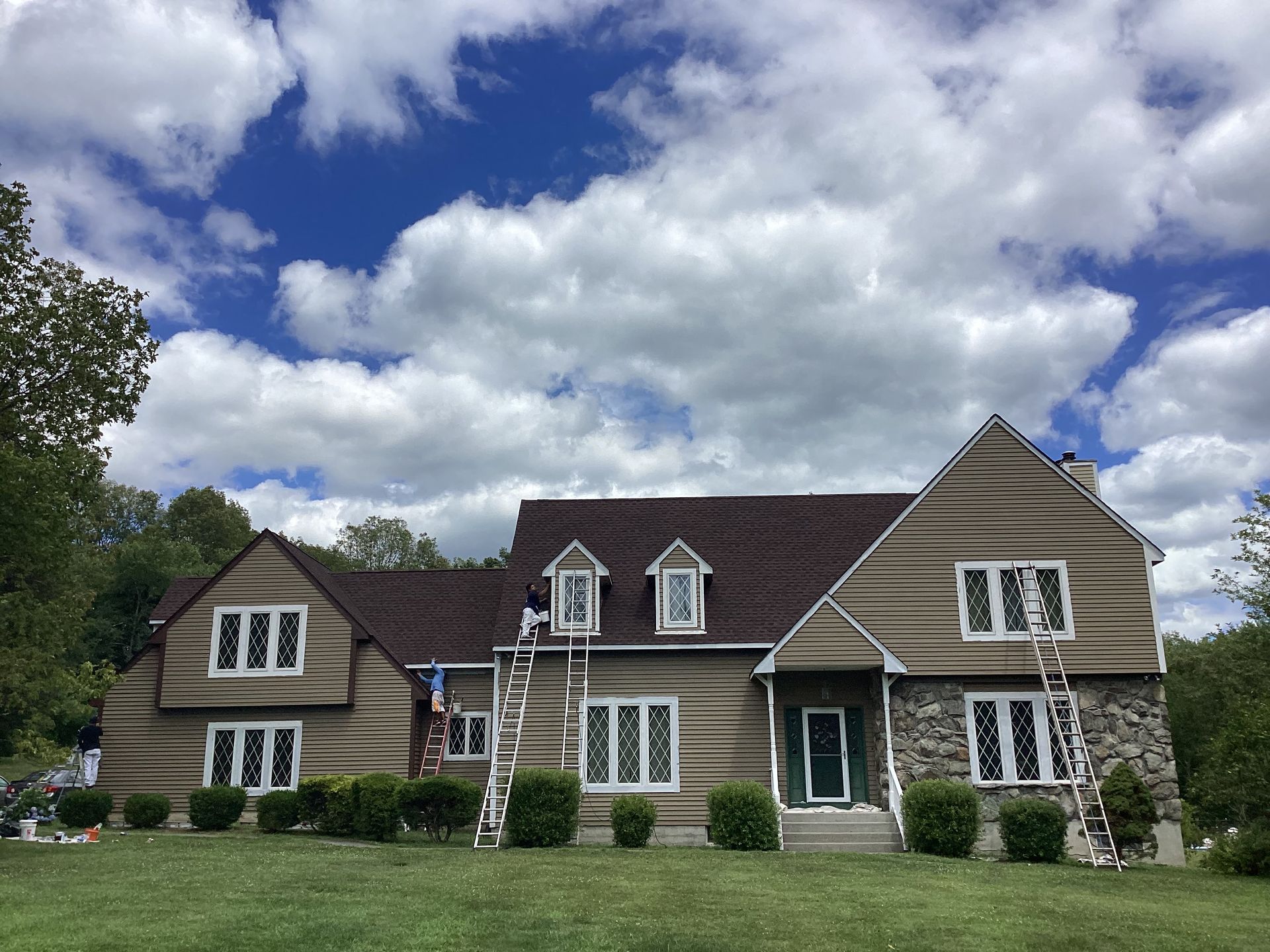 House being painted, brown siding, white trim, green door, ladders, blue sky, sunny day.