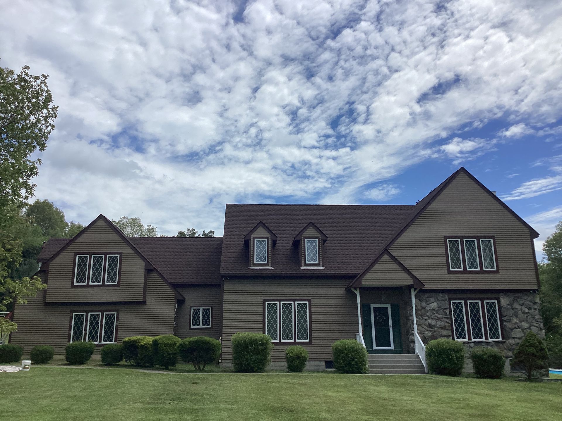 Two-story house with brown siding, stone accents, and brown roof under a partly cloudy sky.