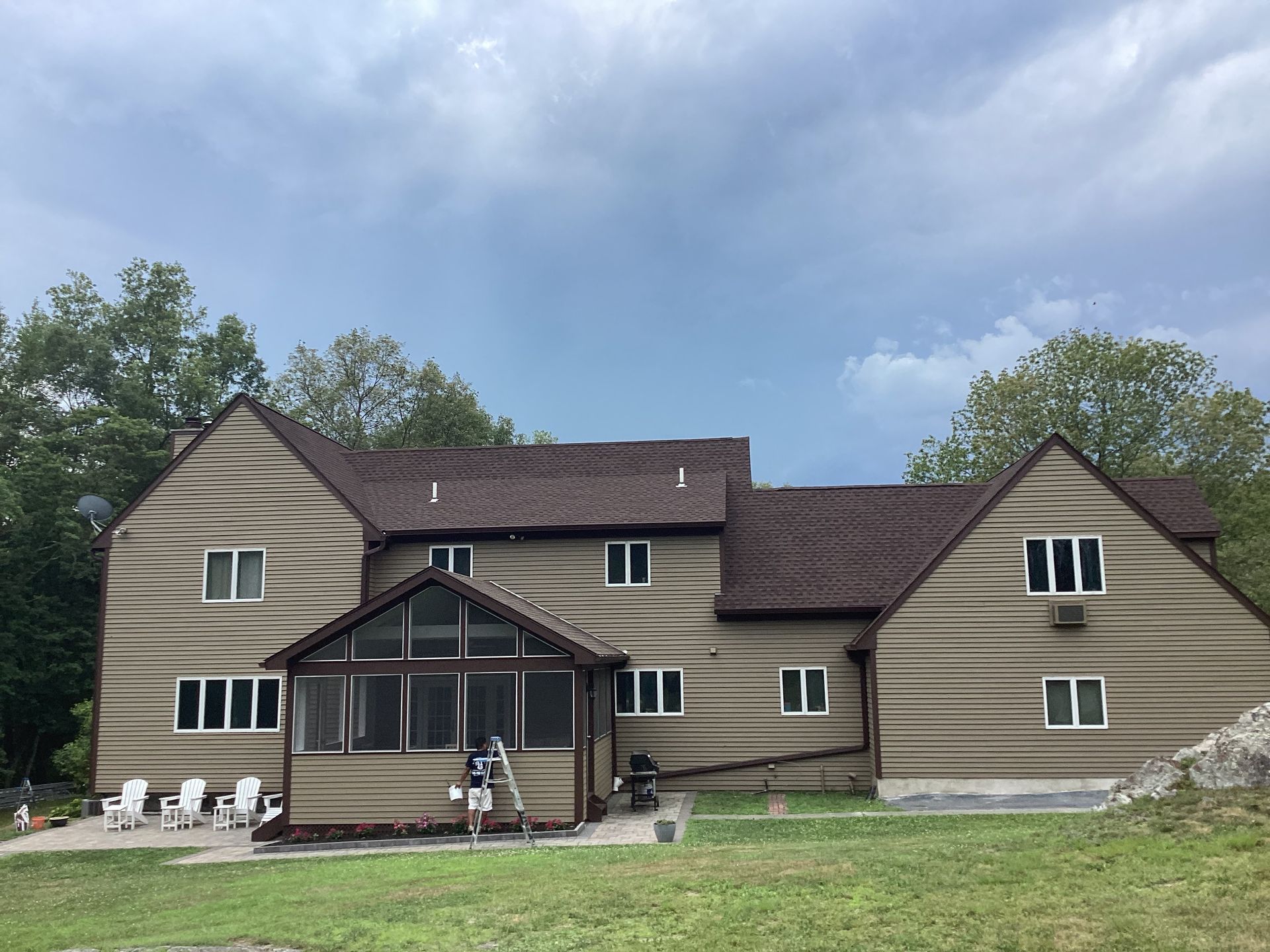 Two-story beige house with brown roof, screened porch, windows, and outdoor chairs on the lawn.