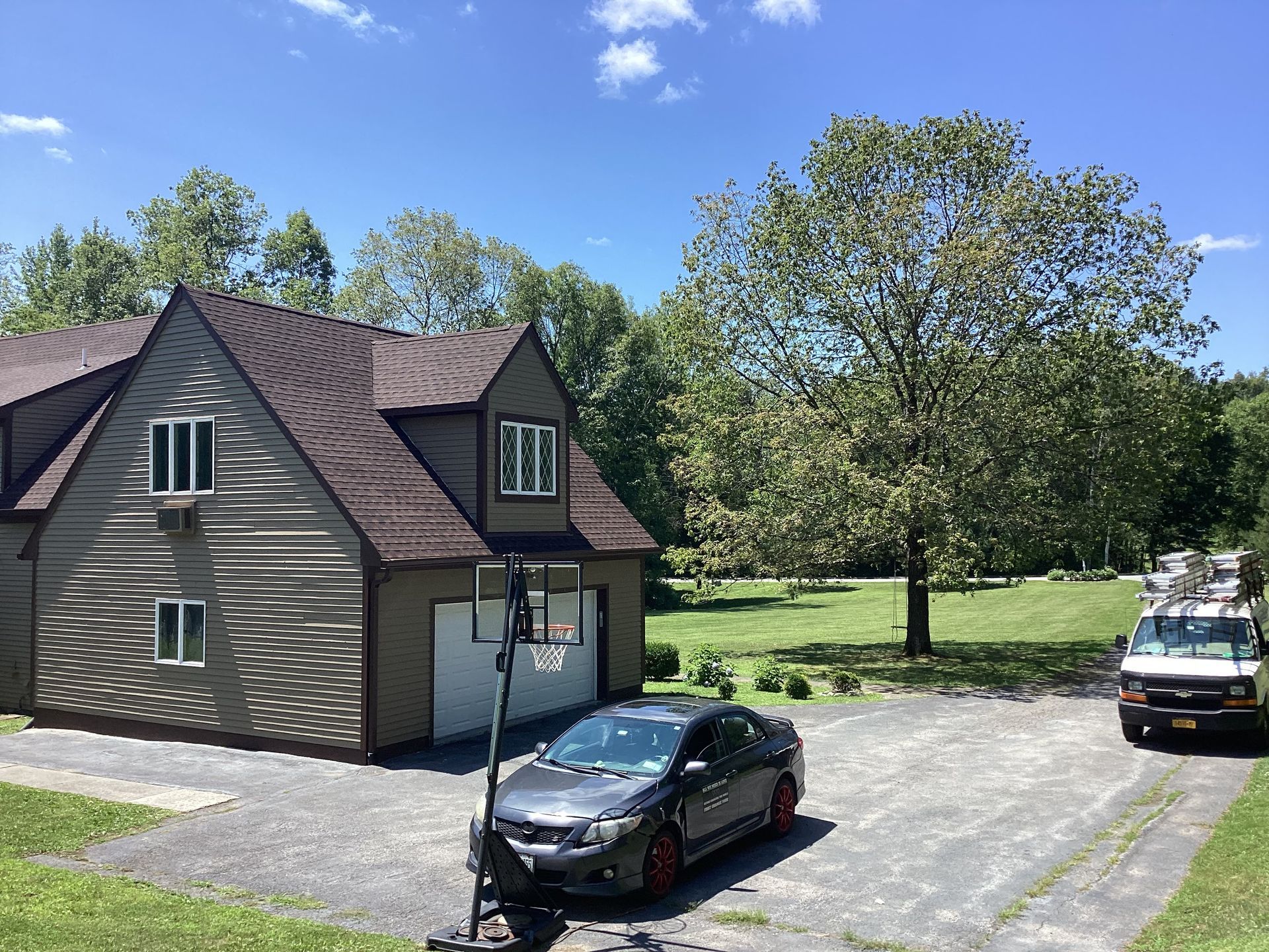 A house with a driveway, car, and basketball hoop on a sunny day. A van is driving away.