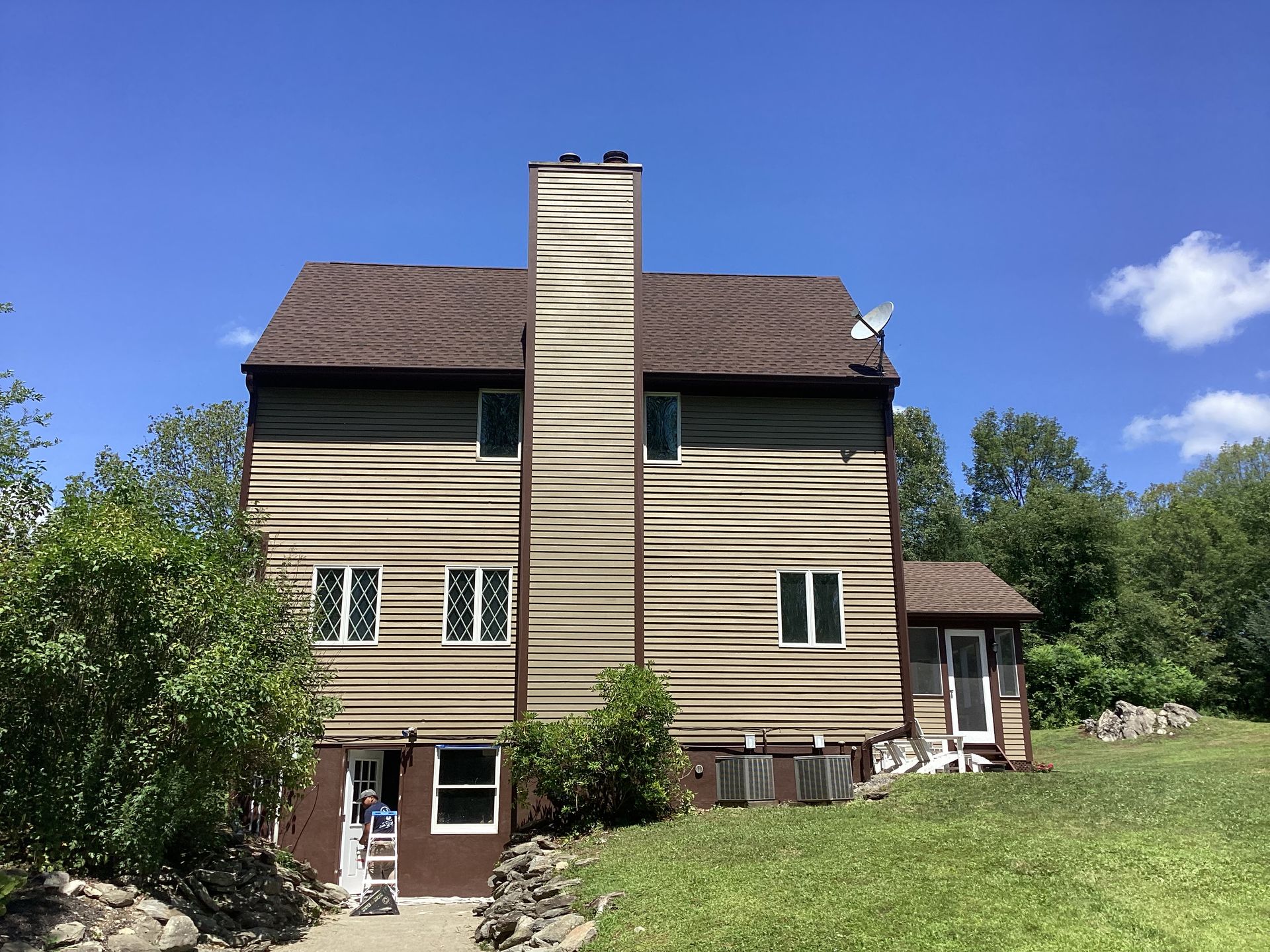 Rear view of a brown wooden house with chimney under a blue sky, surrounded by green trees.