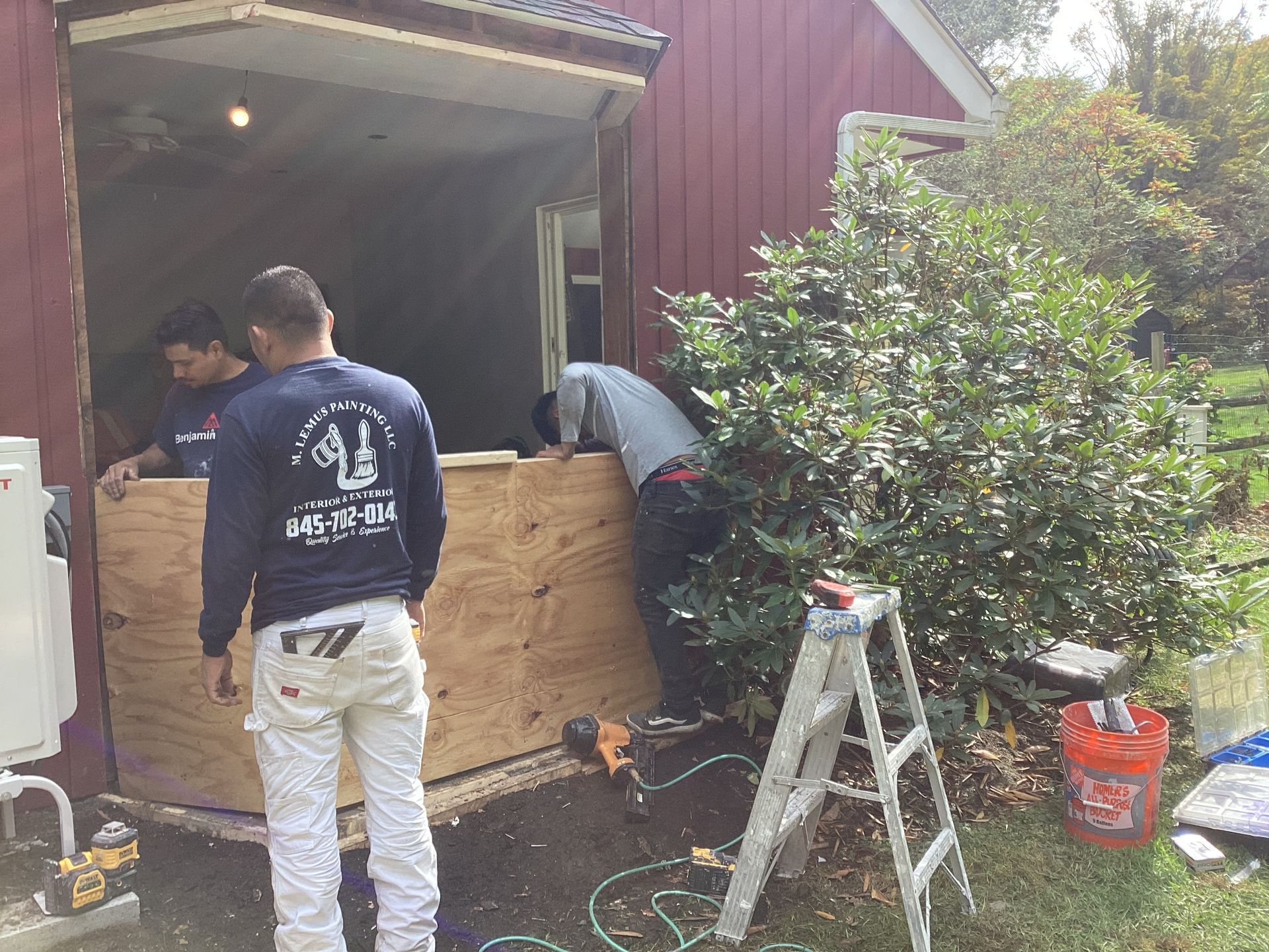 Three men installing plywood into a red building's window frame; a ladder and shrubs are nearby.