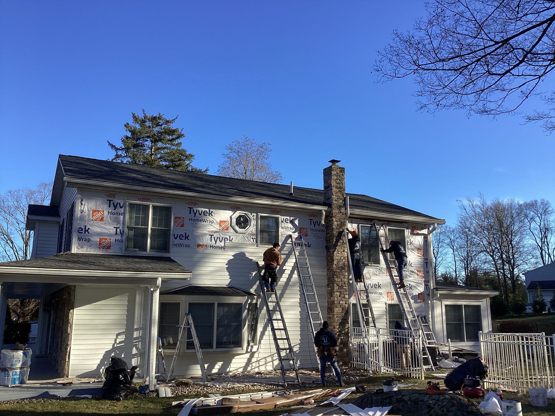 House siding being replaced; workers on ladders, applying new material. Blue sky, sunny day.