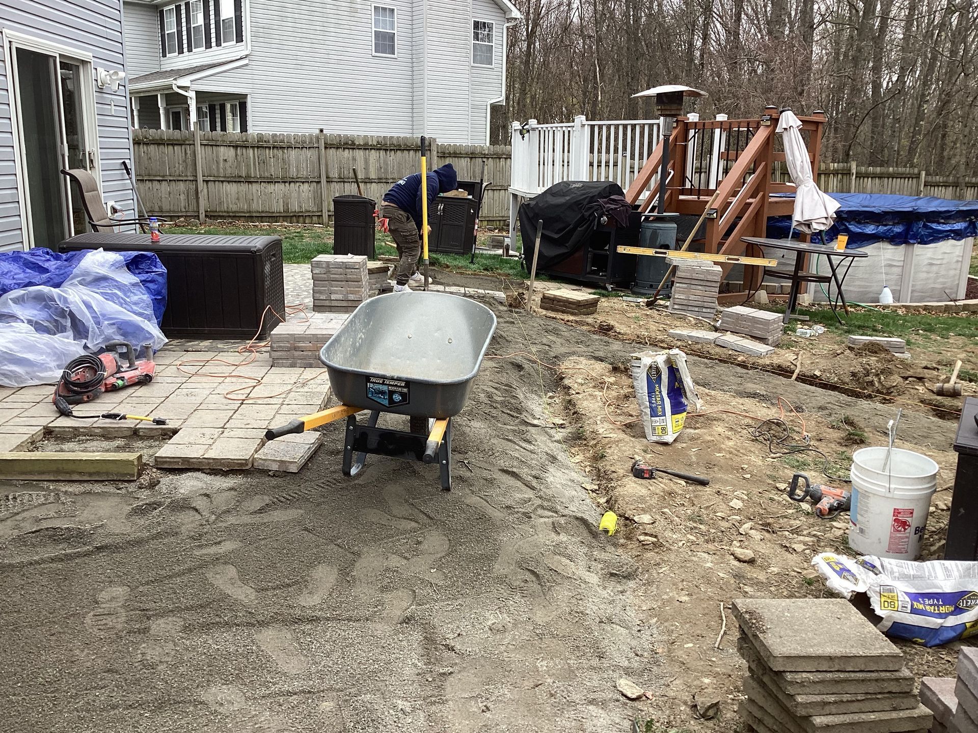 Backyard patio construction: Wheelbarrow, pavers, person working, outdoor setting.