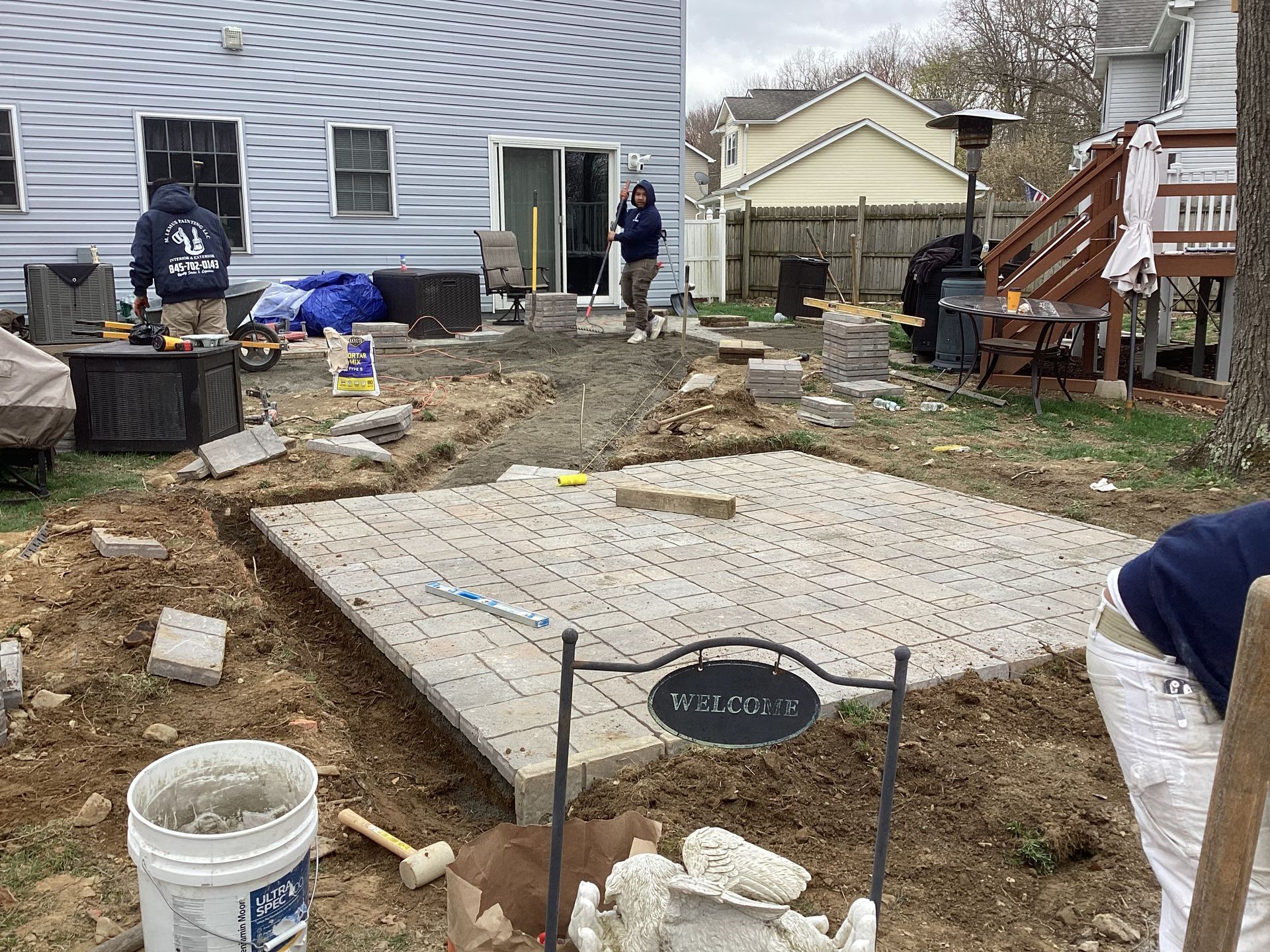Backyard patio construction in progress; workers, pavers, dirt, tools.