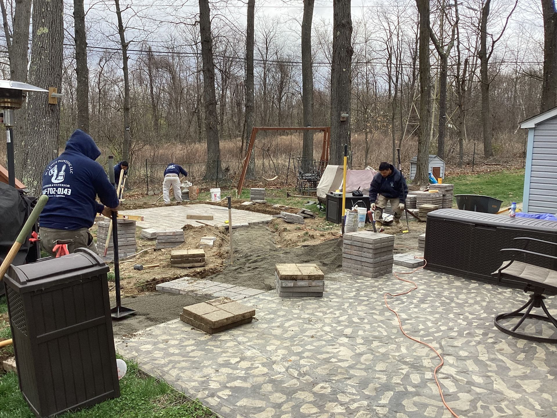 Workers laying pavers in a backyard; trees in the background, overcast day.
