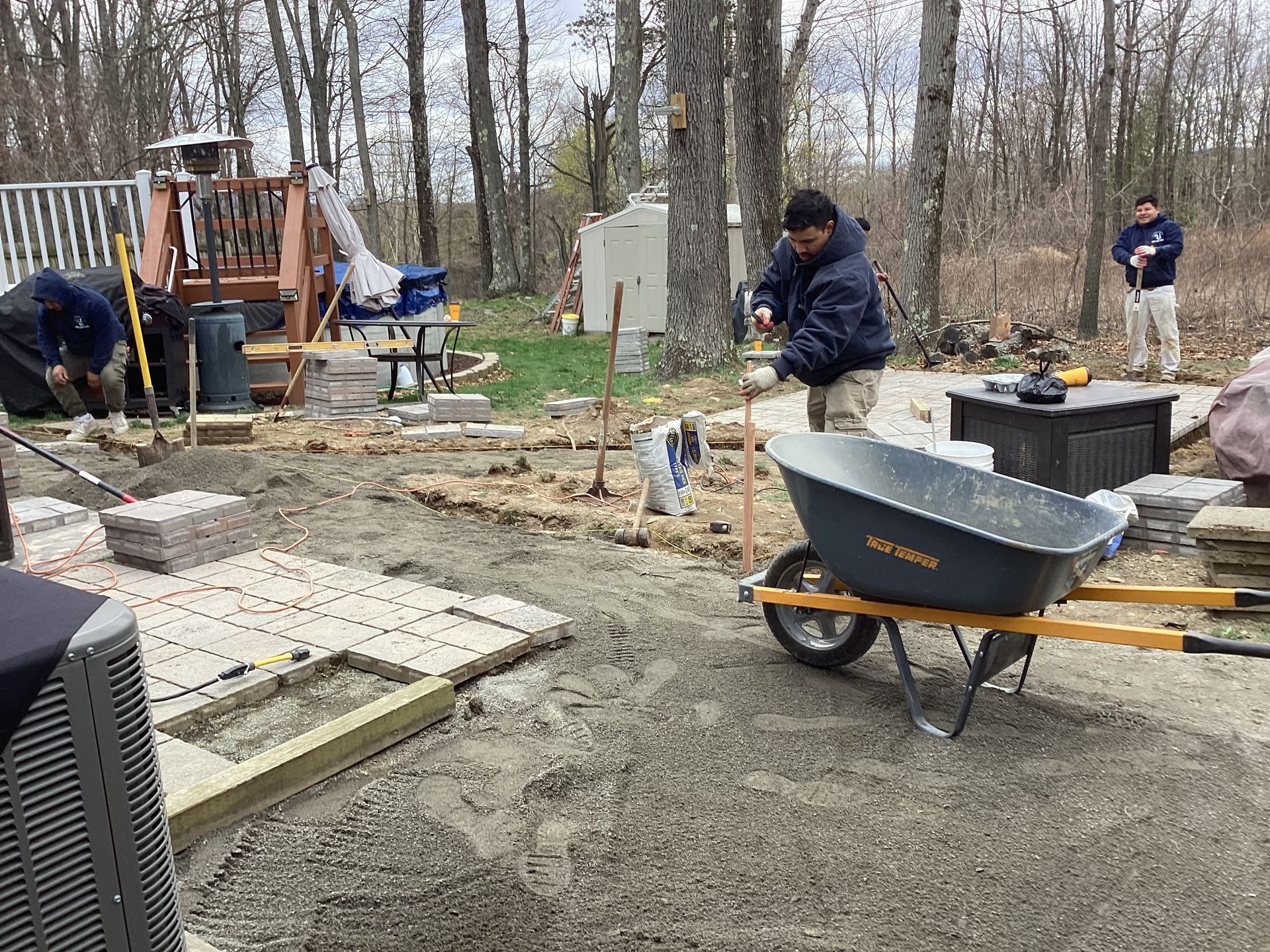 People laying pavers in a backyard. A wheelbarrow, tools, and building materials are present.