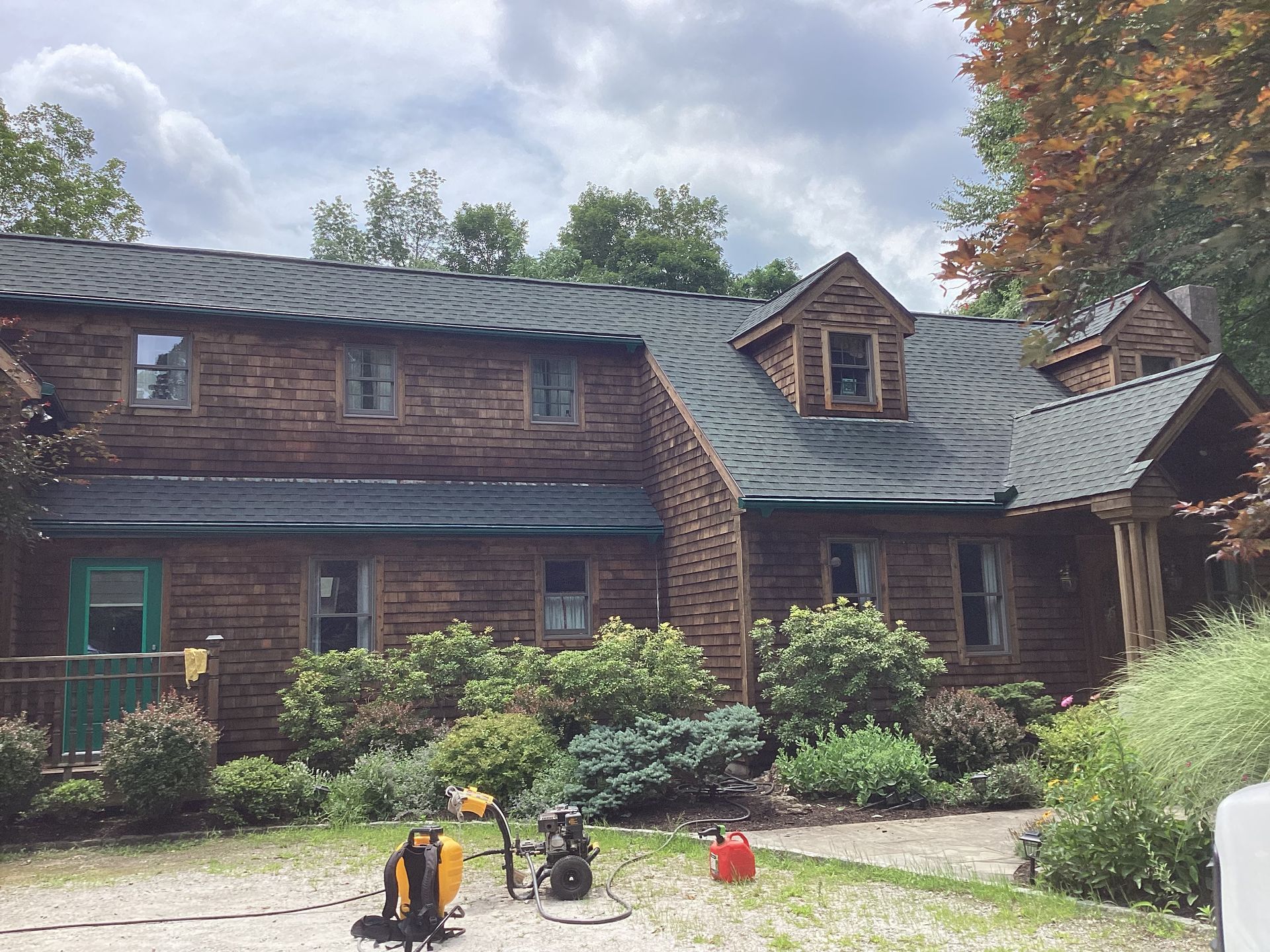 Brown brick house with a dark green metal roof, bushes, and tools in the yard.