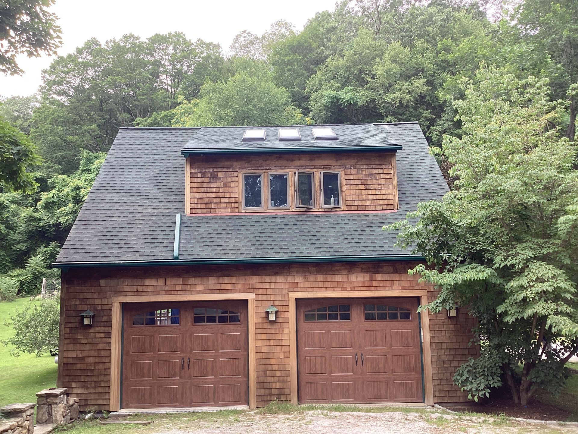 Two-story wooden garage with brown doors and windows, a dark shingle roof, and trees in the background.