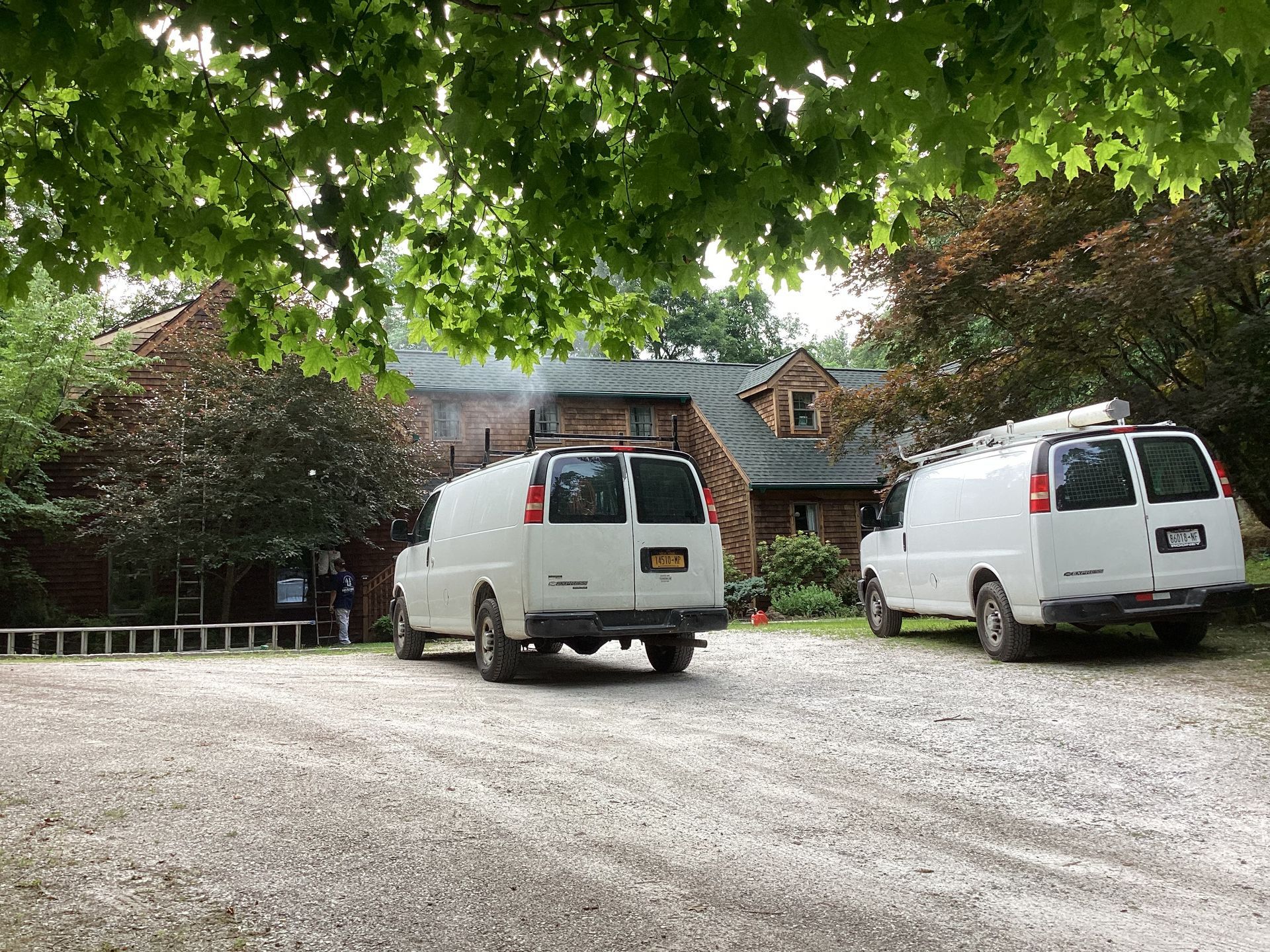 Two white vans parked on a gravel driveway in front of a brick building, under leafy trees.