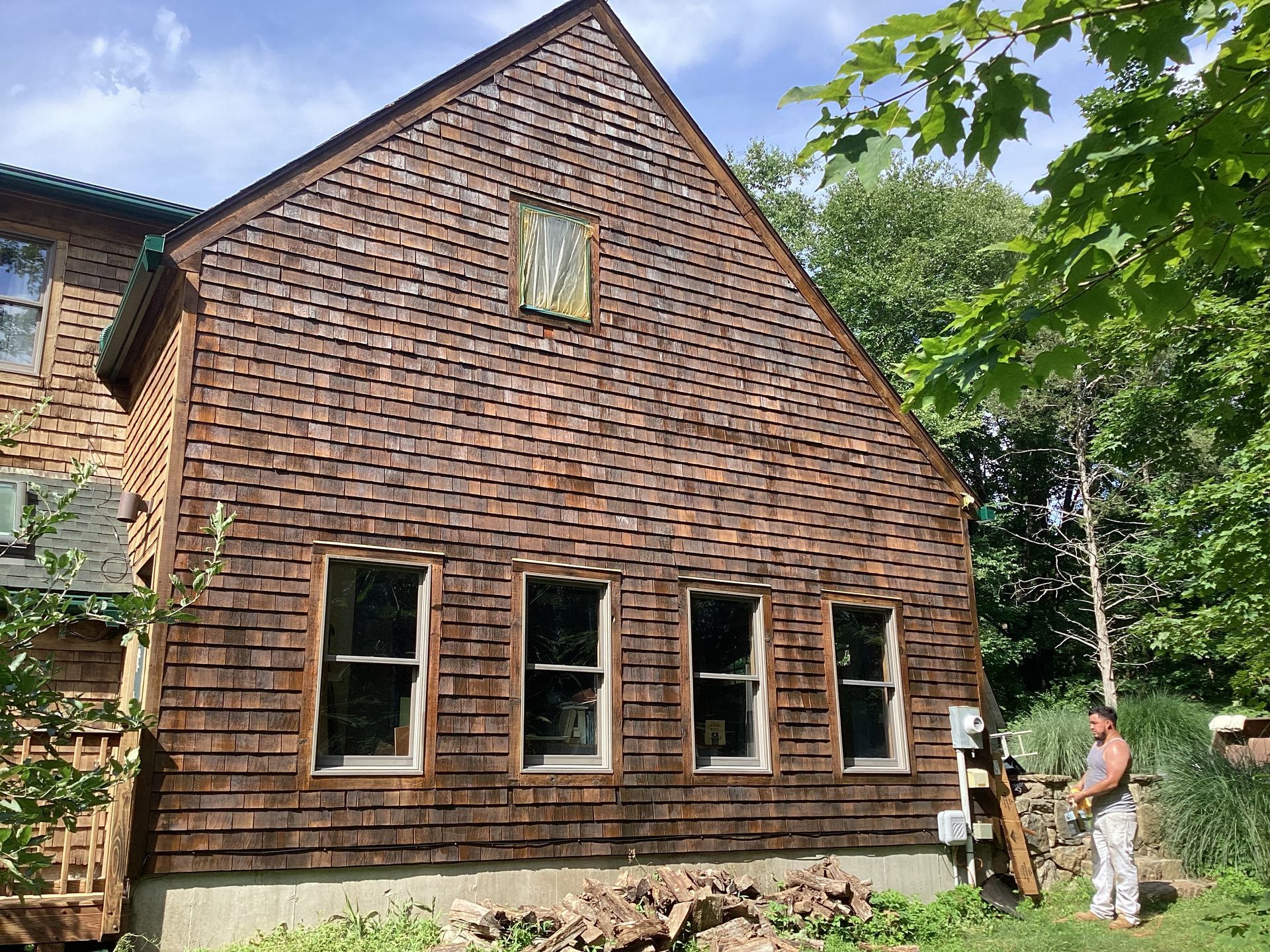 A man stands by a shingled house with several windows and overgrown greenery.