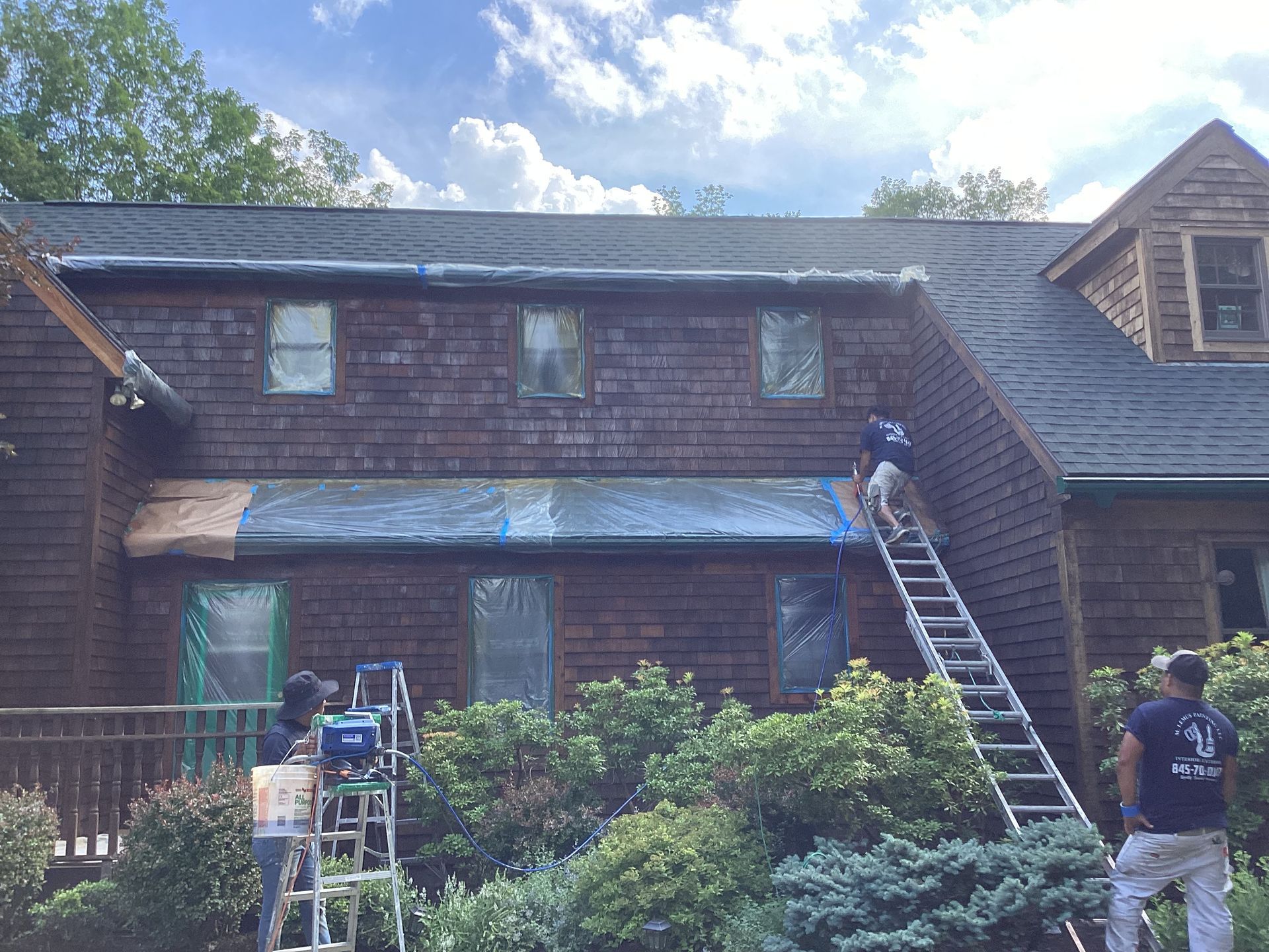 Workers painting a brown shingled house on a sunny day. Men on ladders and ground, with protective coverings.