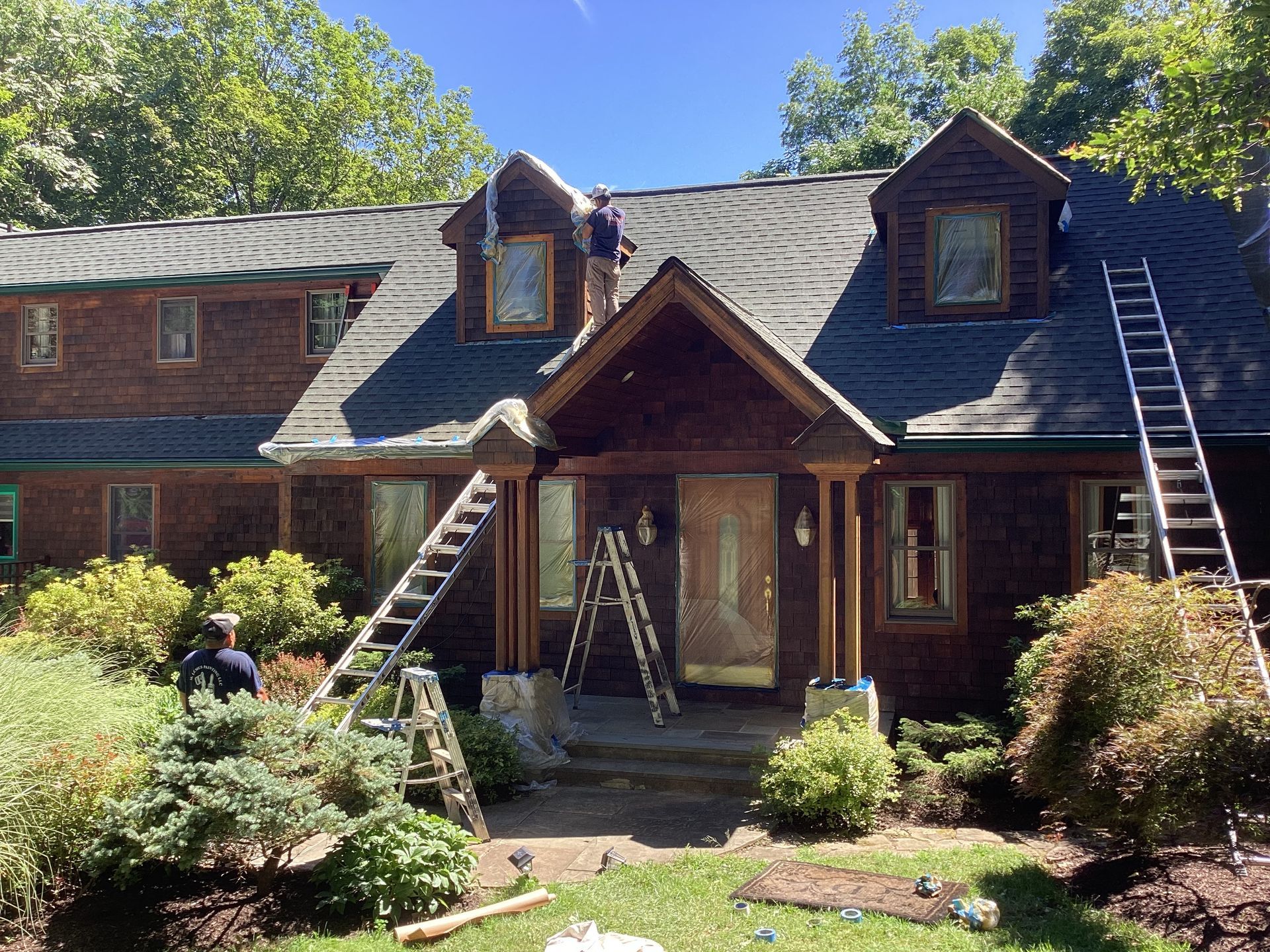 Workers on roof and ground painting a dark wood house with ladders, surrounded by greenery.