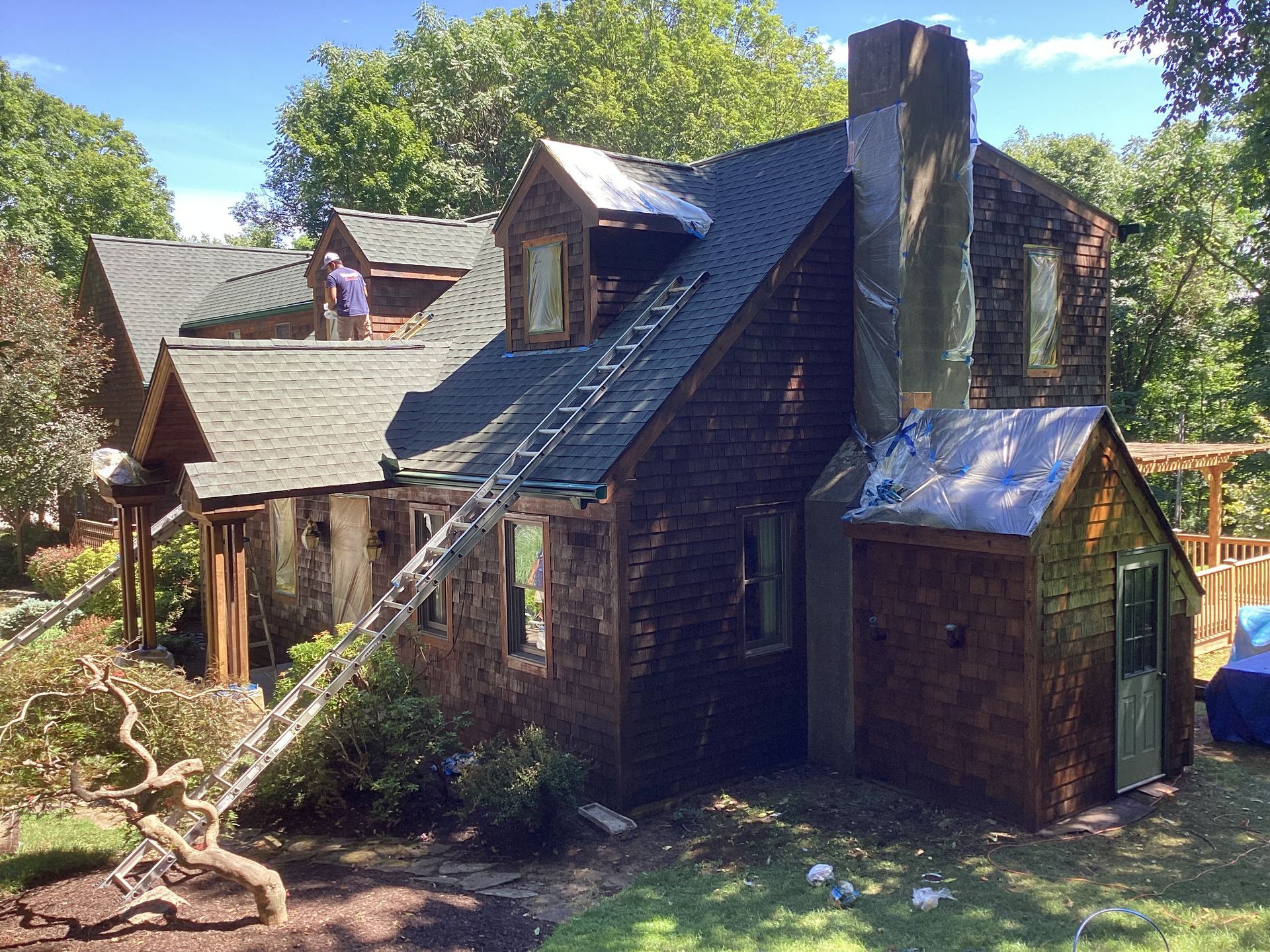 House under construction; roof partially removed, worker on roof, ladder propped up, cedar siding, sunny day.