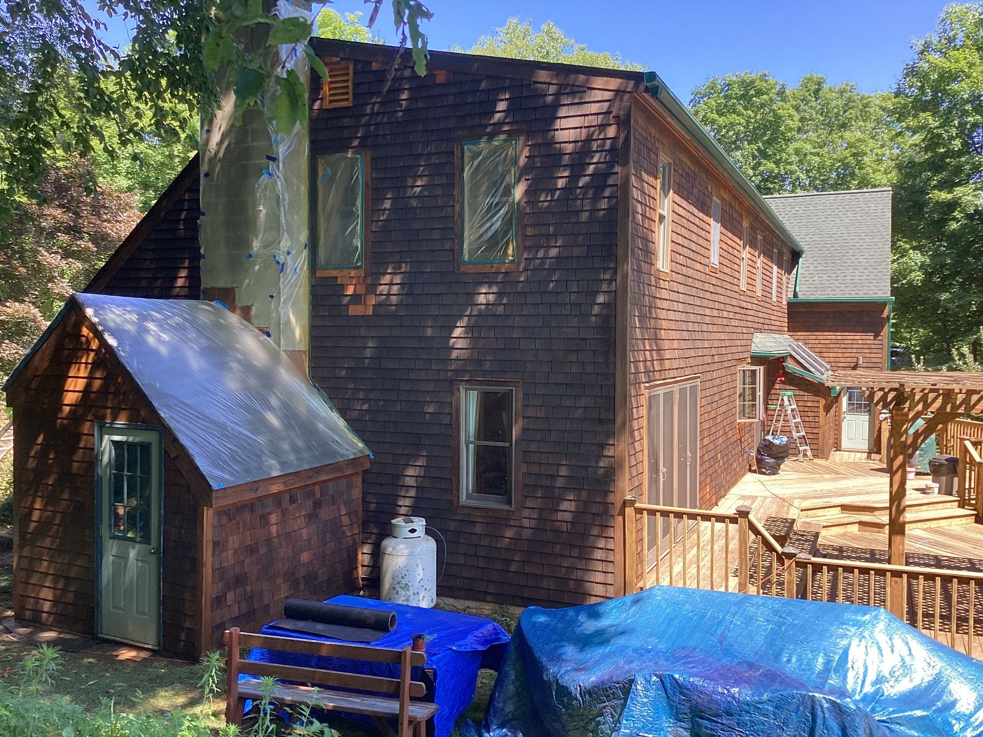 Two-story brown house with a shed, deck, and tarp-covered objects in a wooded setting.