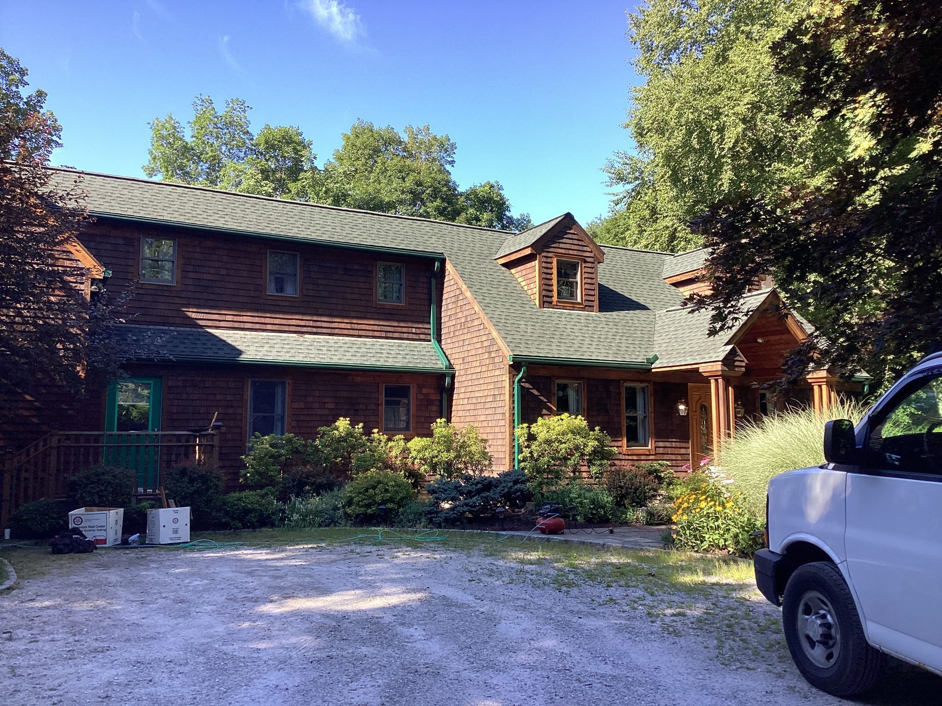 Two-story log cabin house with green roof and trim, surrounded by greenery and a white van in the driveway.
