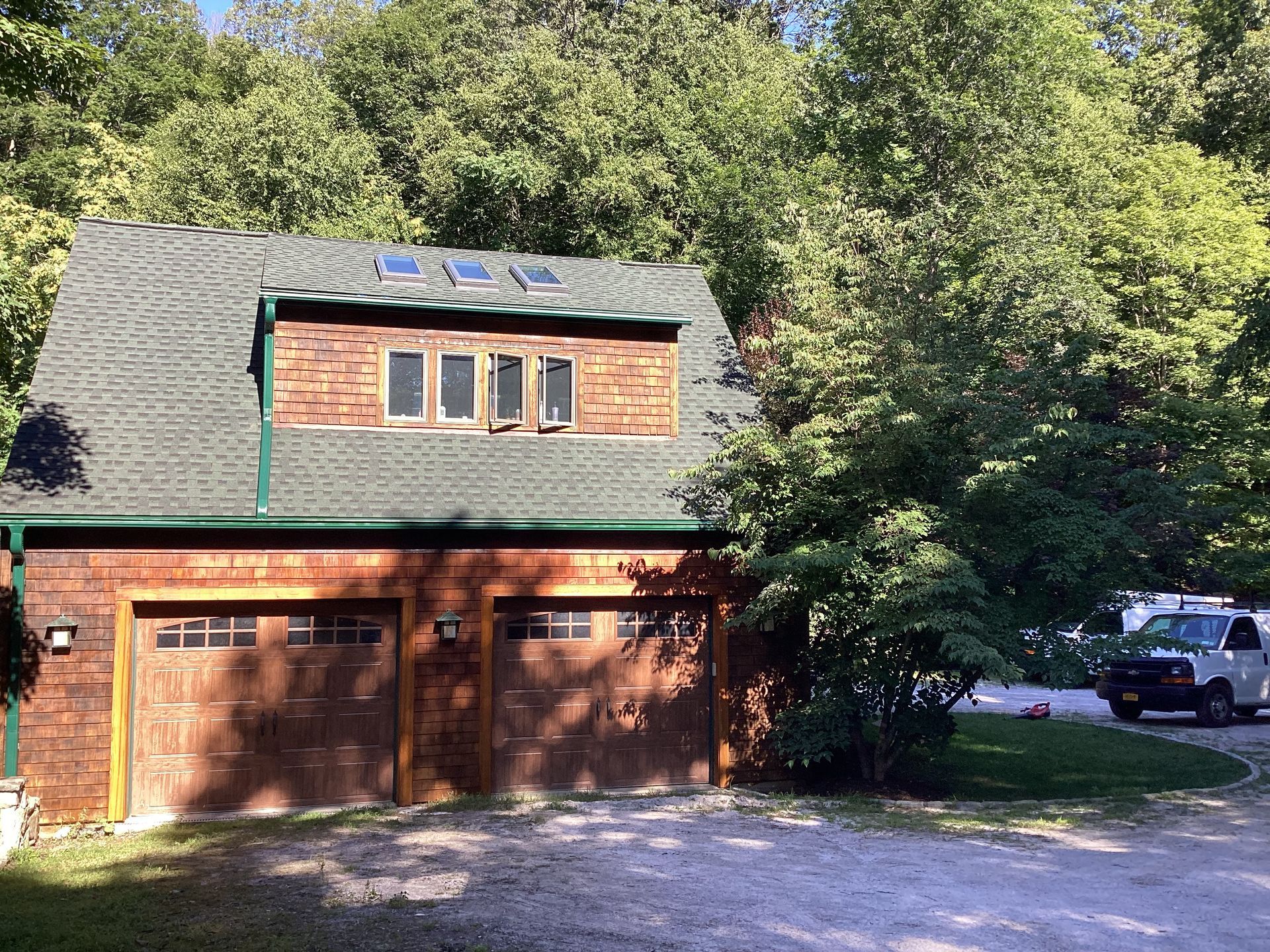 Garage with brown doors, brick facade, green roof, and dormer with windows surrounded by trees.