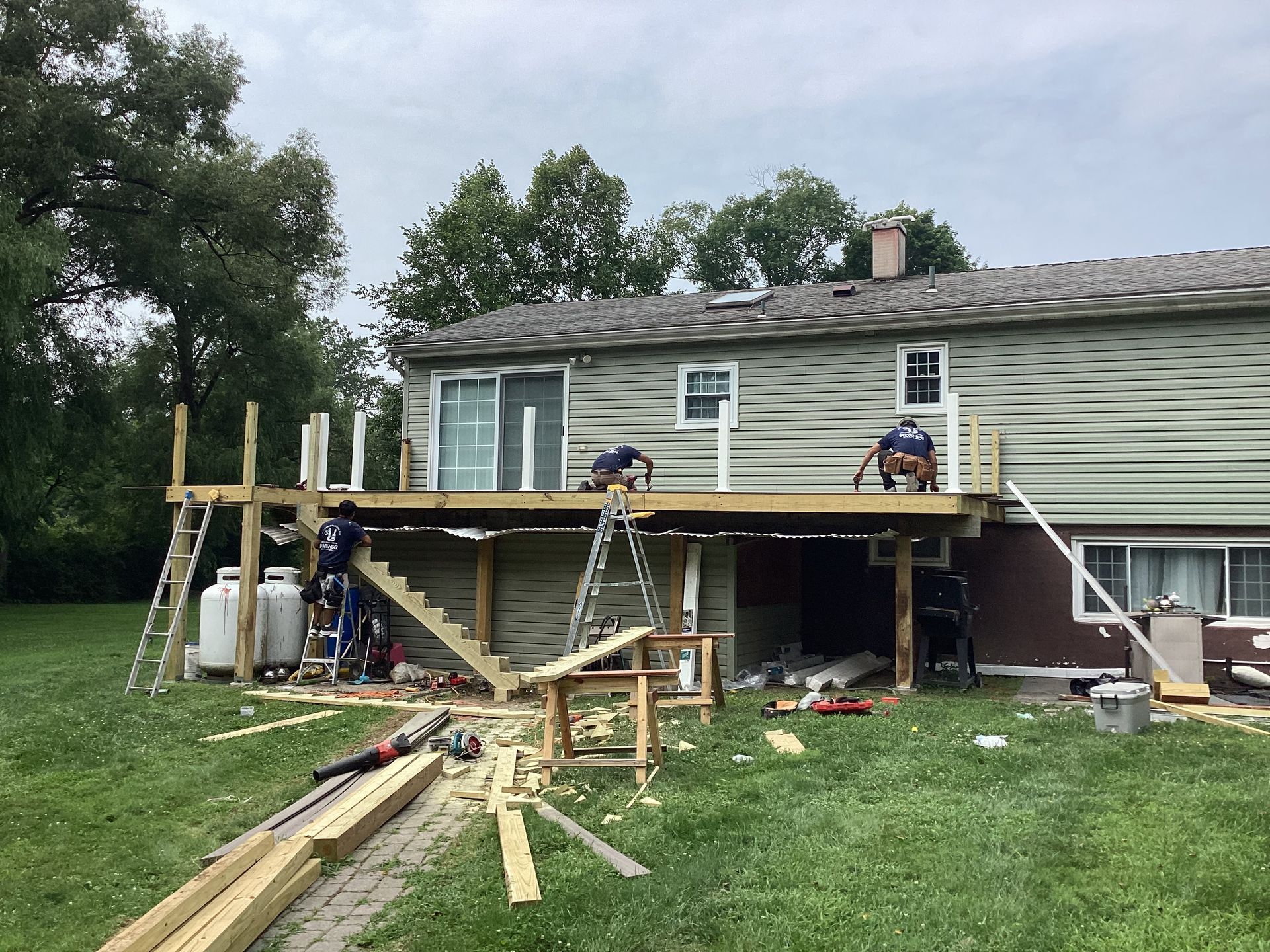 Construction workers building a wooden deck attached to a house with green siding on a cloudy day.