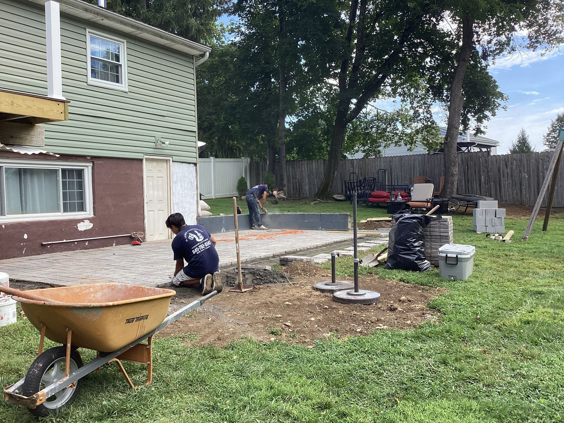 Workers laying pavers in backyard, with a wheelbarrow, and building foundation. Green grass, cloudy sky.