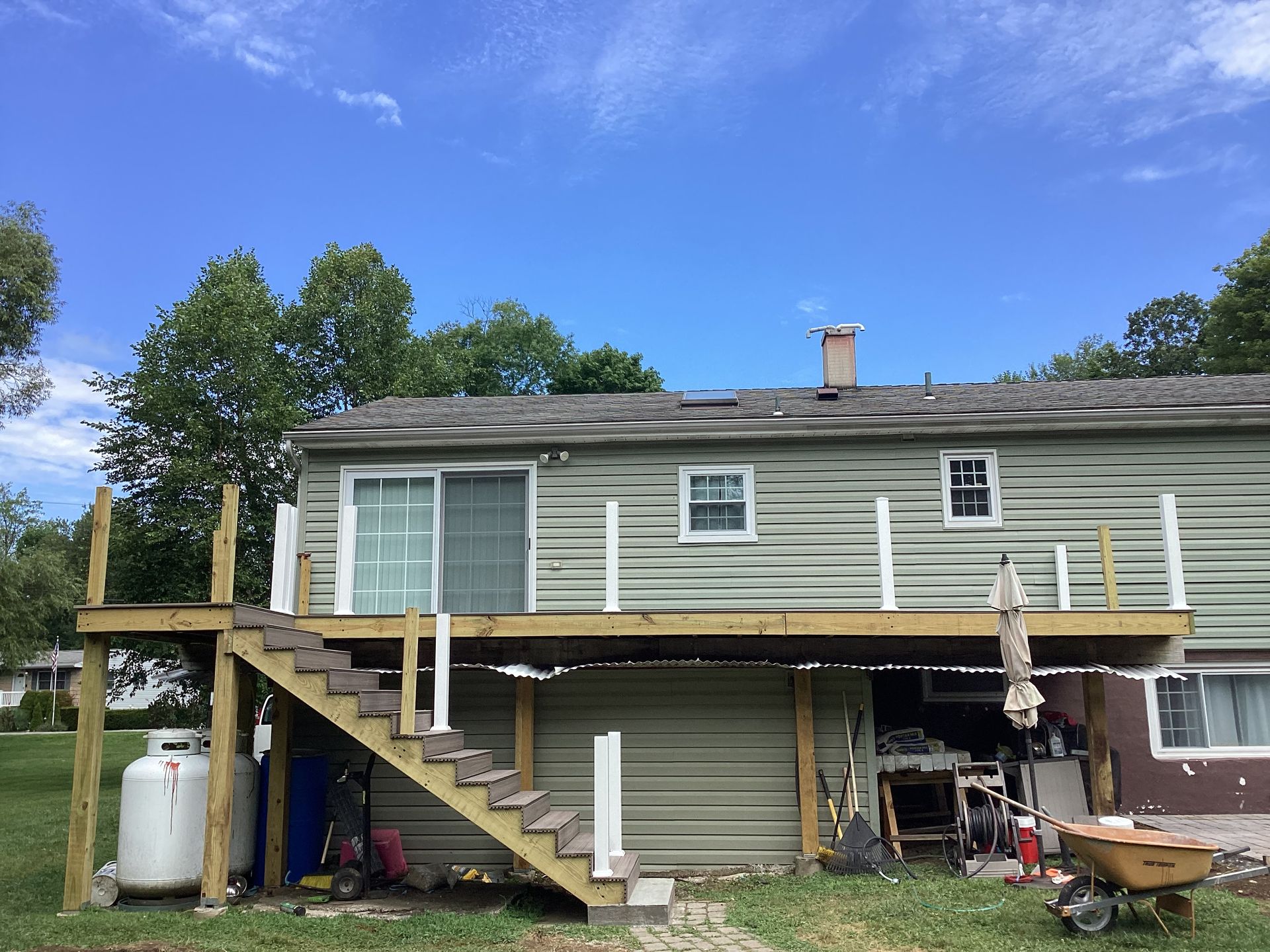 Back of house with a wooden deck and stairs, with blue sky and trees in the background.