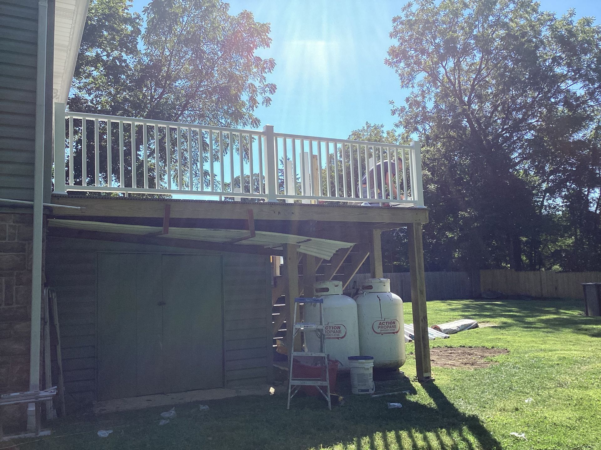 A raised wooden deck with white railing over a shed, propane tanks visible. Sunlight above, green grass below.