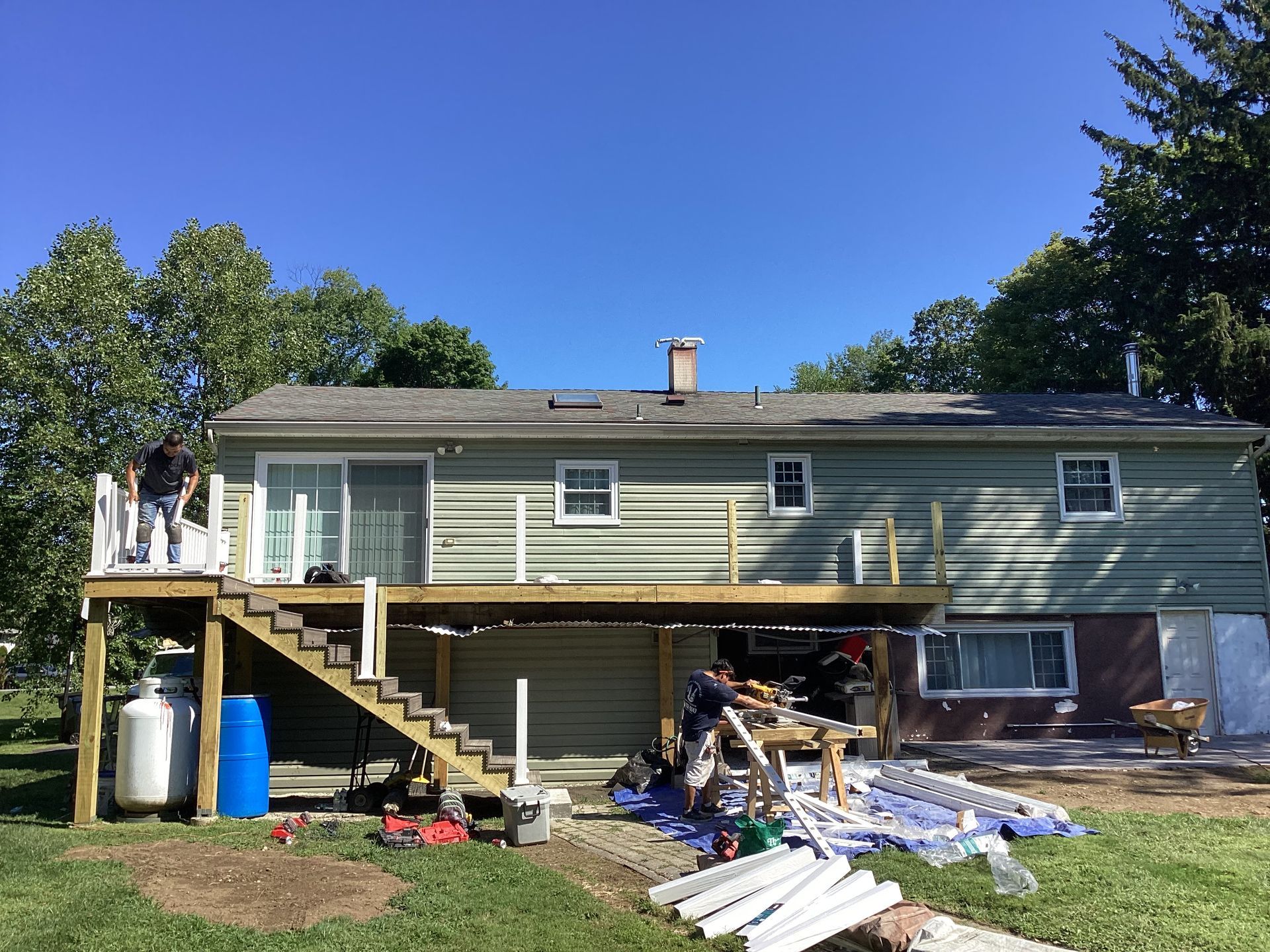 Rear view of a house with a partially built deck; two men working on the deck, blue sky background.