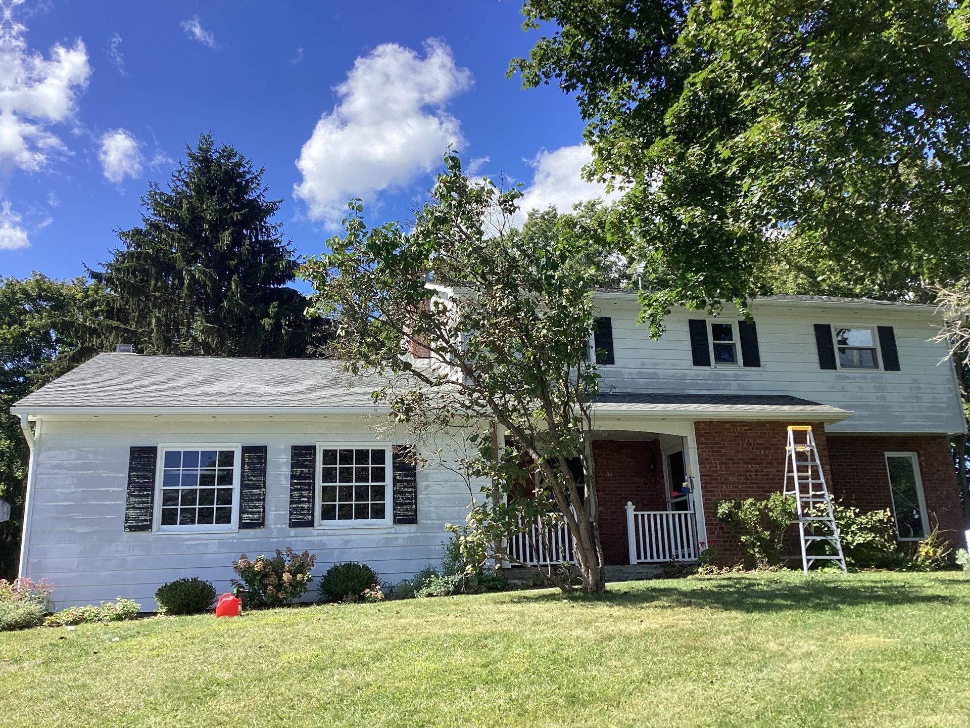 Two-story white house with black shutters, brick entry, and a ladder, trees and grass.