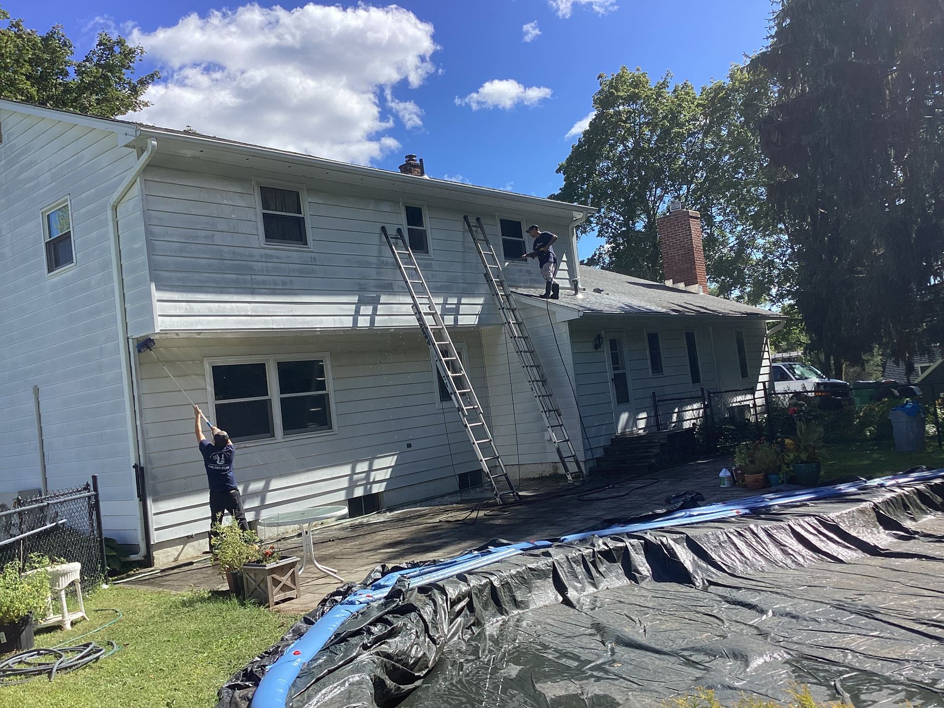 Workers on ladders near a house, pool in the foreground, cloudy sky.