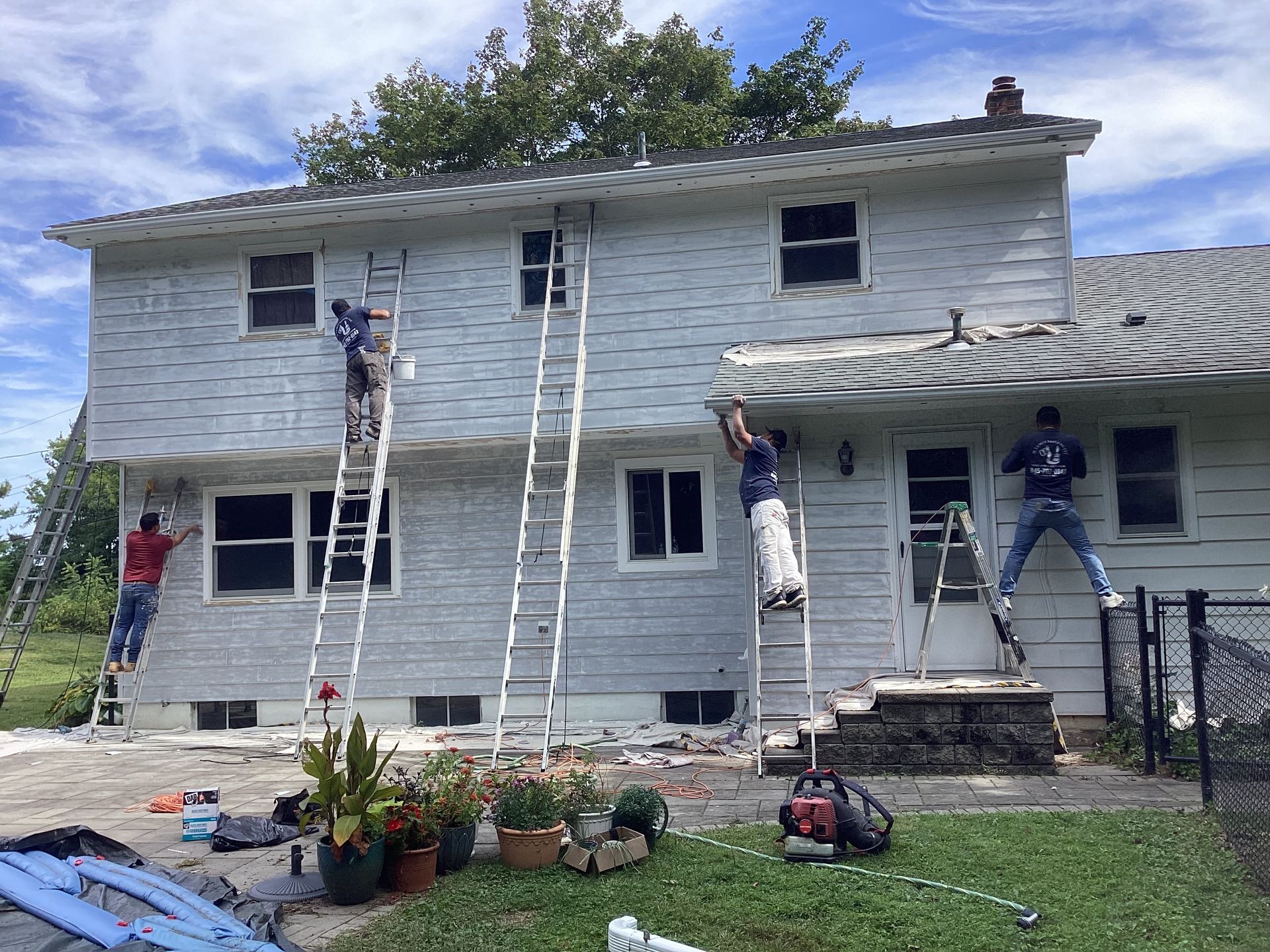 Workers painting a two-story house. Several people on ladders. Gray siding, white trim. Sunny day.