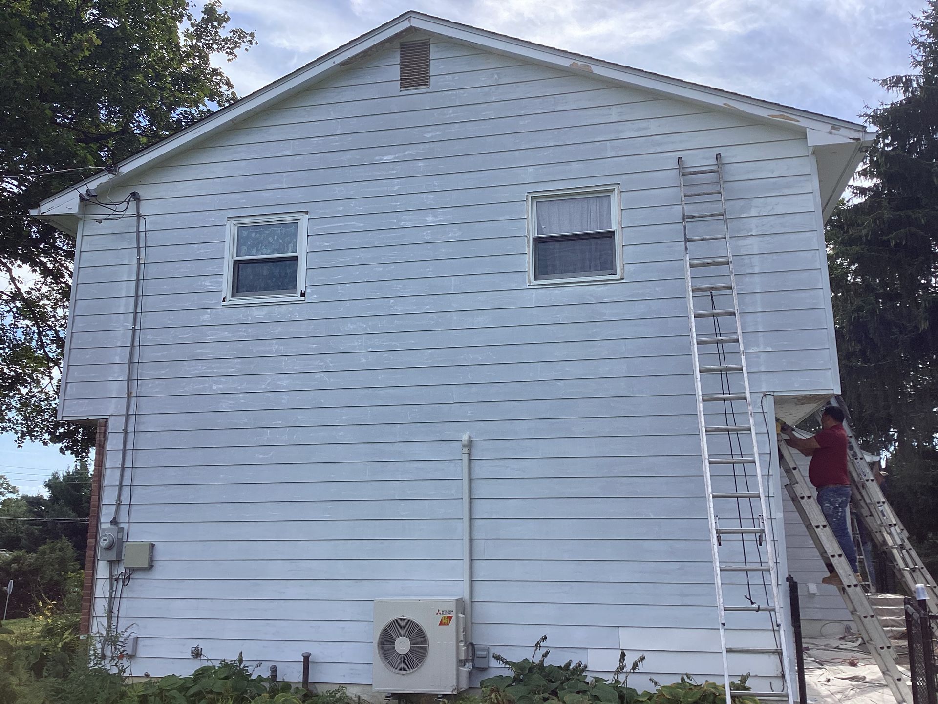 Side of a two-story house with a person on a ladder by a door. White siding, two windows, and an air conditioner are visible.