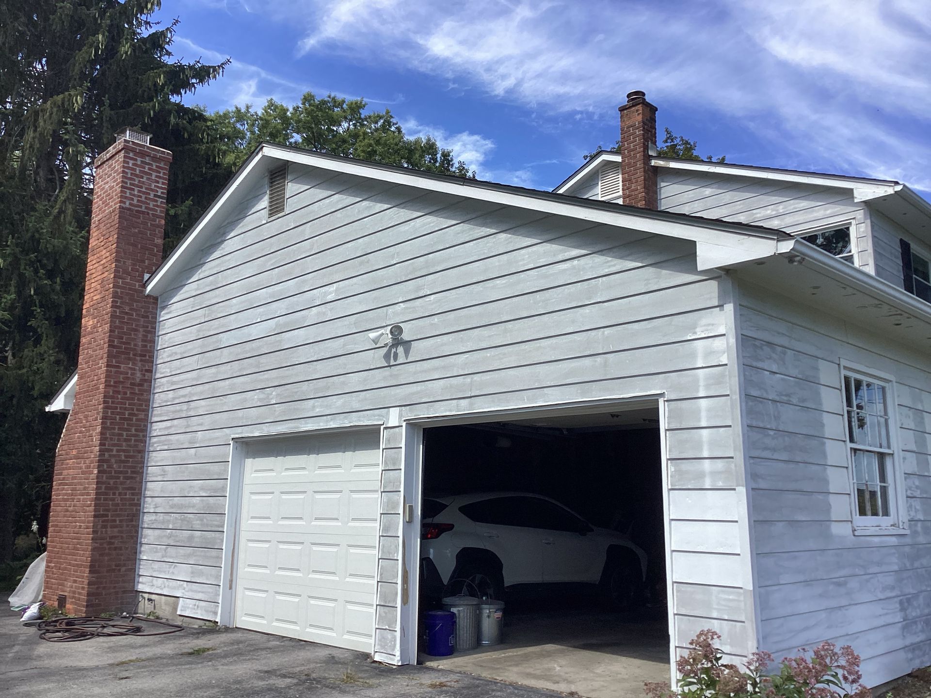 Garage with white door, gray siding, brick chimney, and a white car inside on a sunny day.