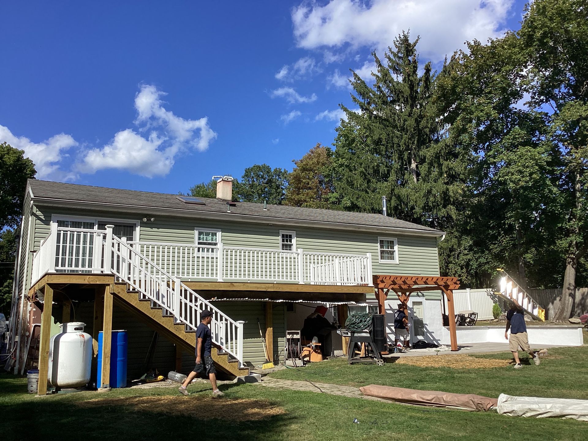 Backyard with two-story house, wooden deck, and pergola. Several people are working under a blue sky.