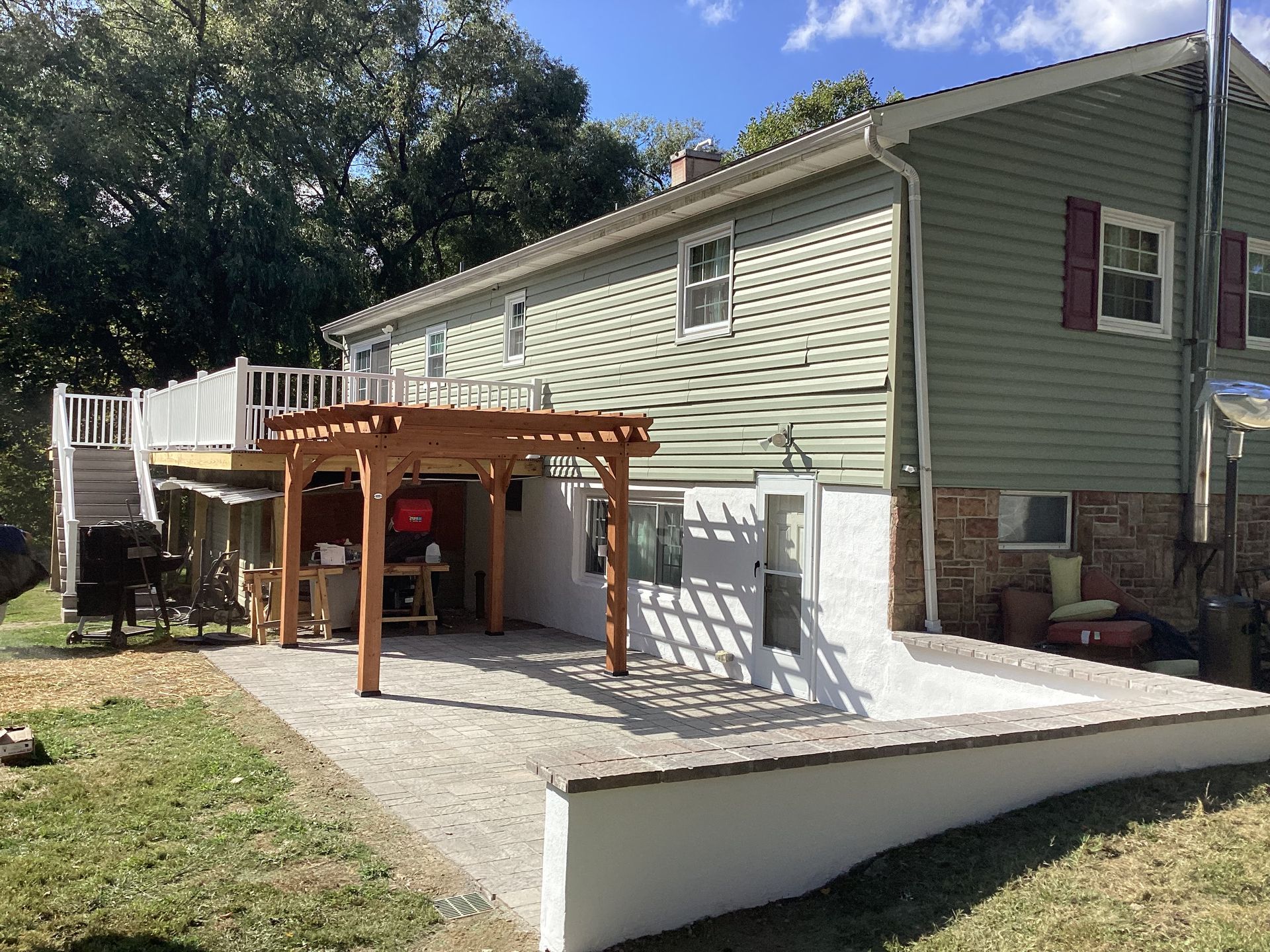 Back of a two-story house with a deck, pergola, and stone patio. Green siding, brown pergola, white deck railing.