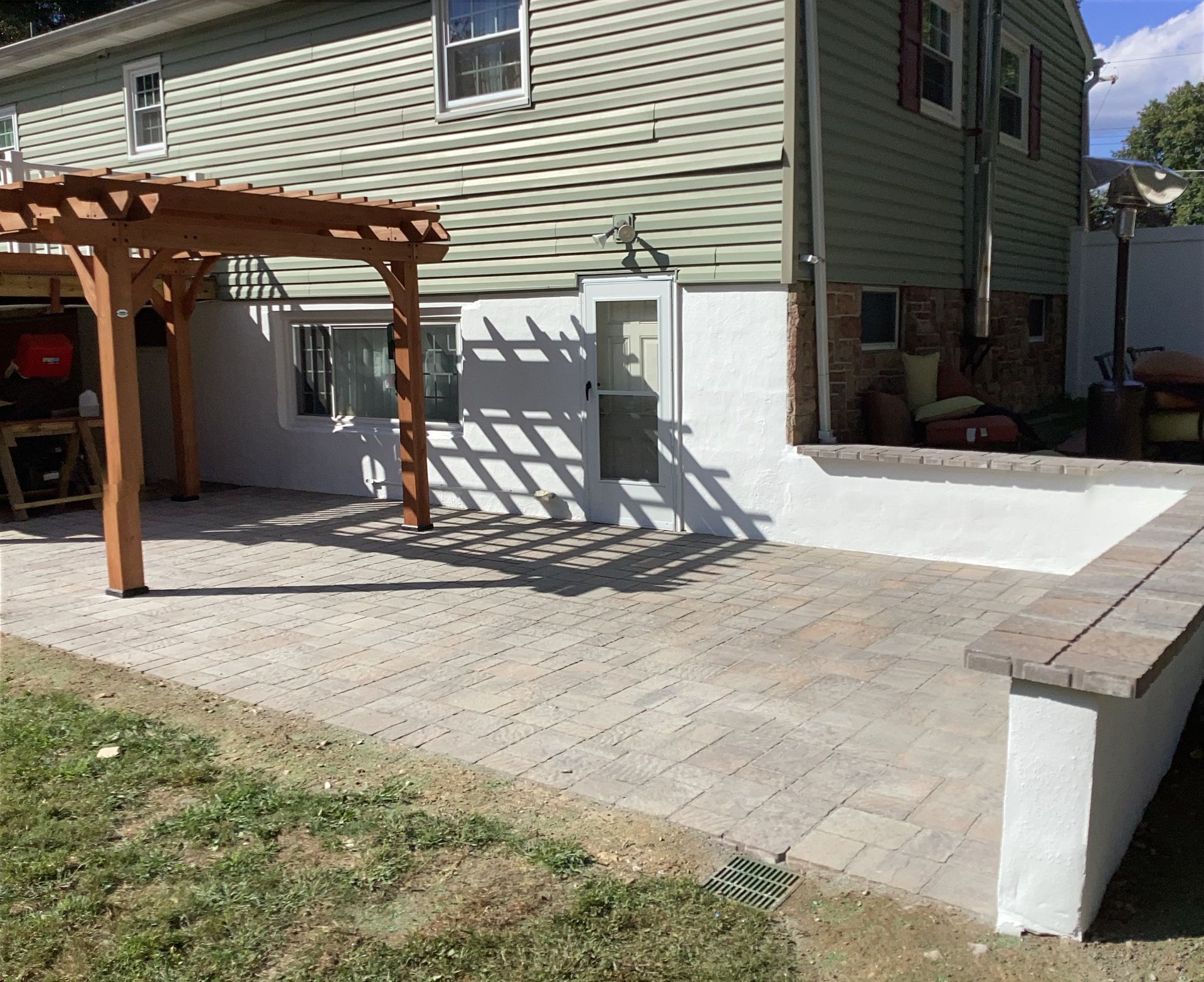 Backyard patio with pergola and low retaining wall; green siding and lawn.