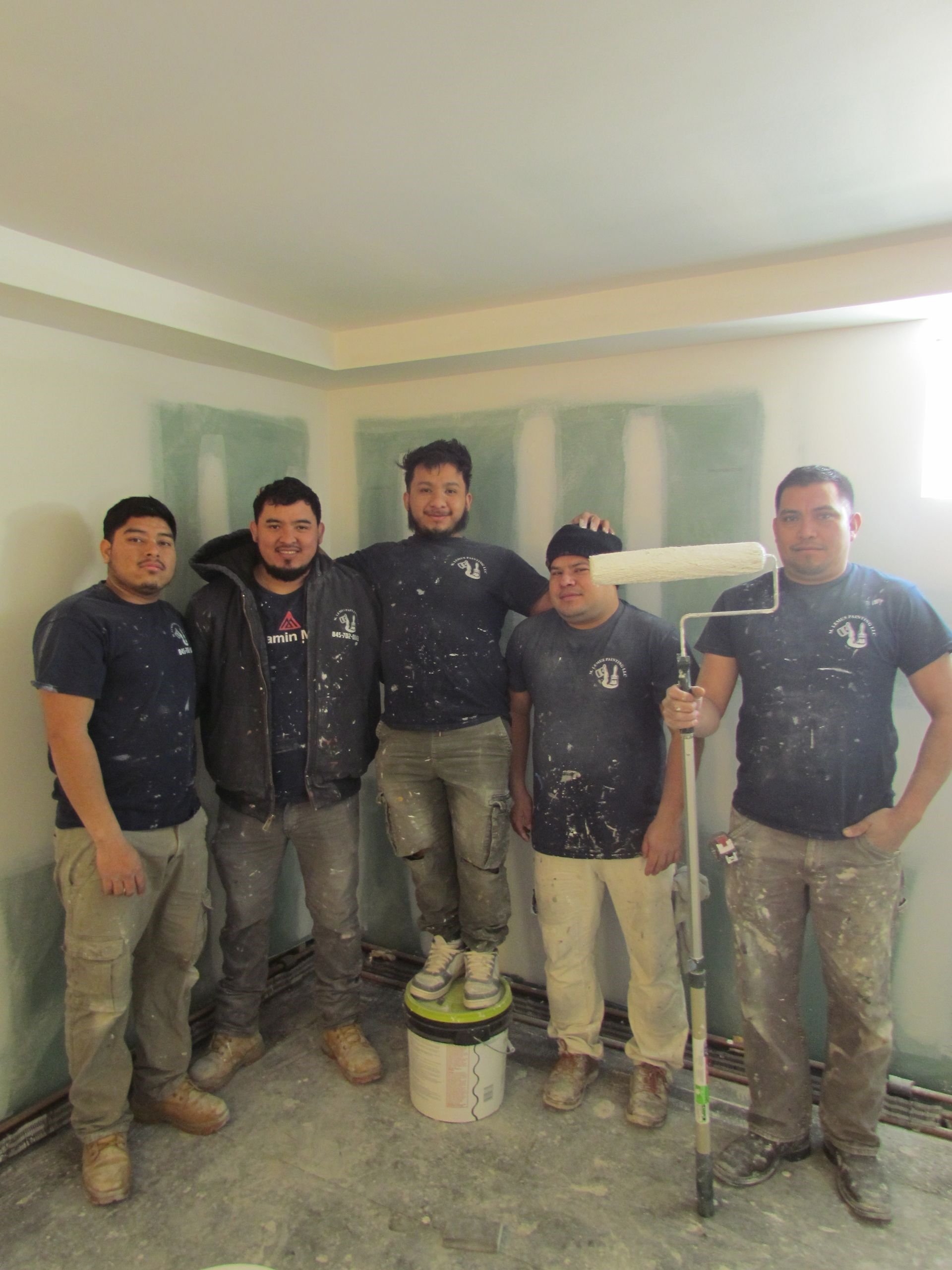 Five people in work clothes stand in a room, paint rollers and buckets present, posing for a photo.