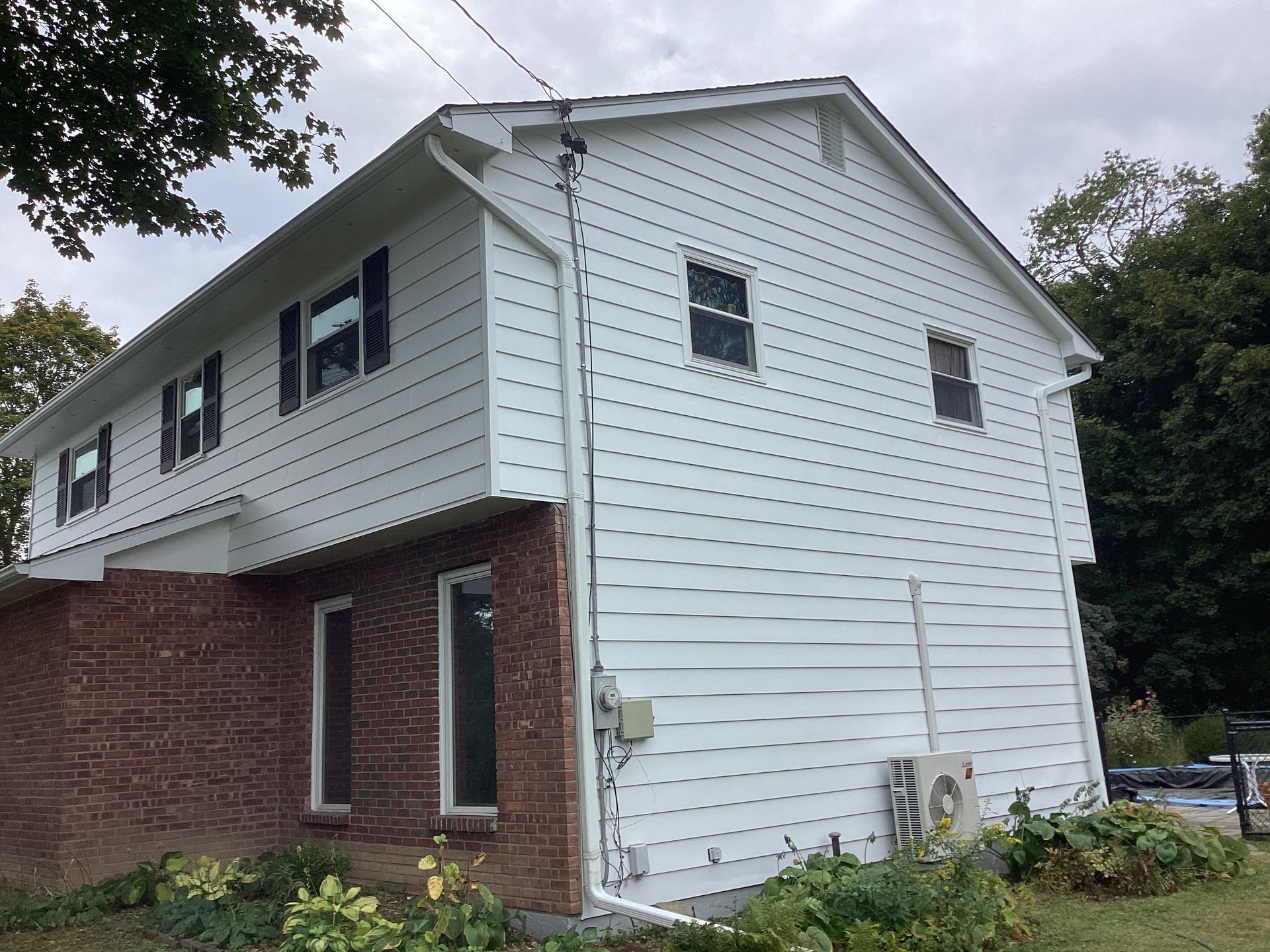 Two-story white house with black shutters, brick facade, and a cloudy sky background.