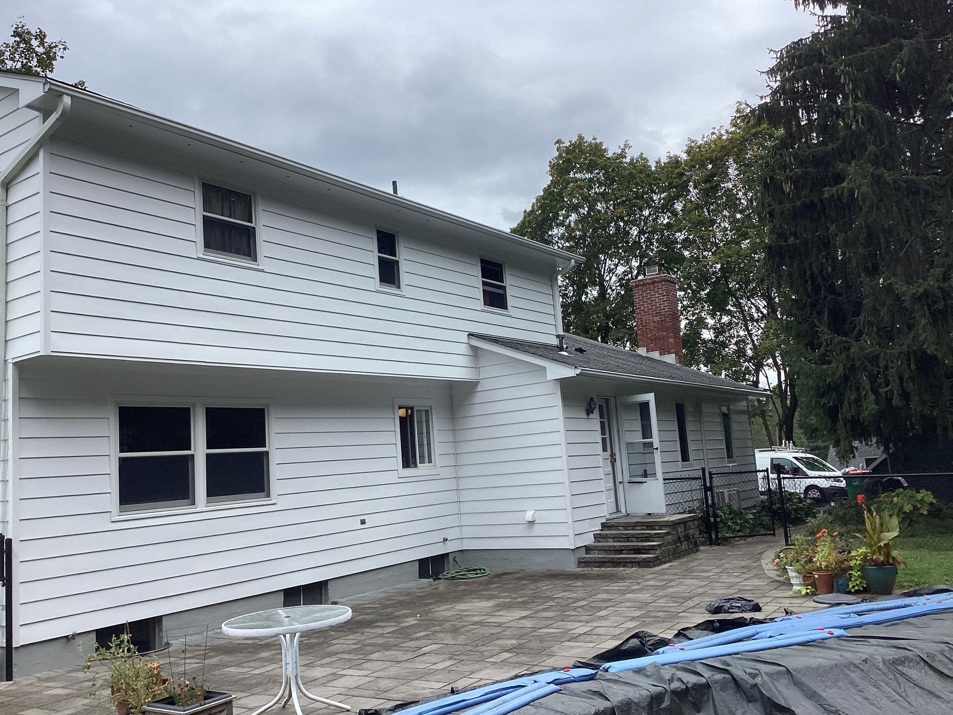 White two-story house with dark windows, gray roof, and concrete patio, under a cloudy sky.