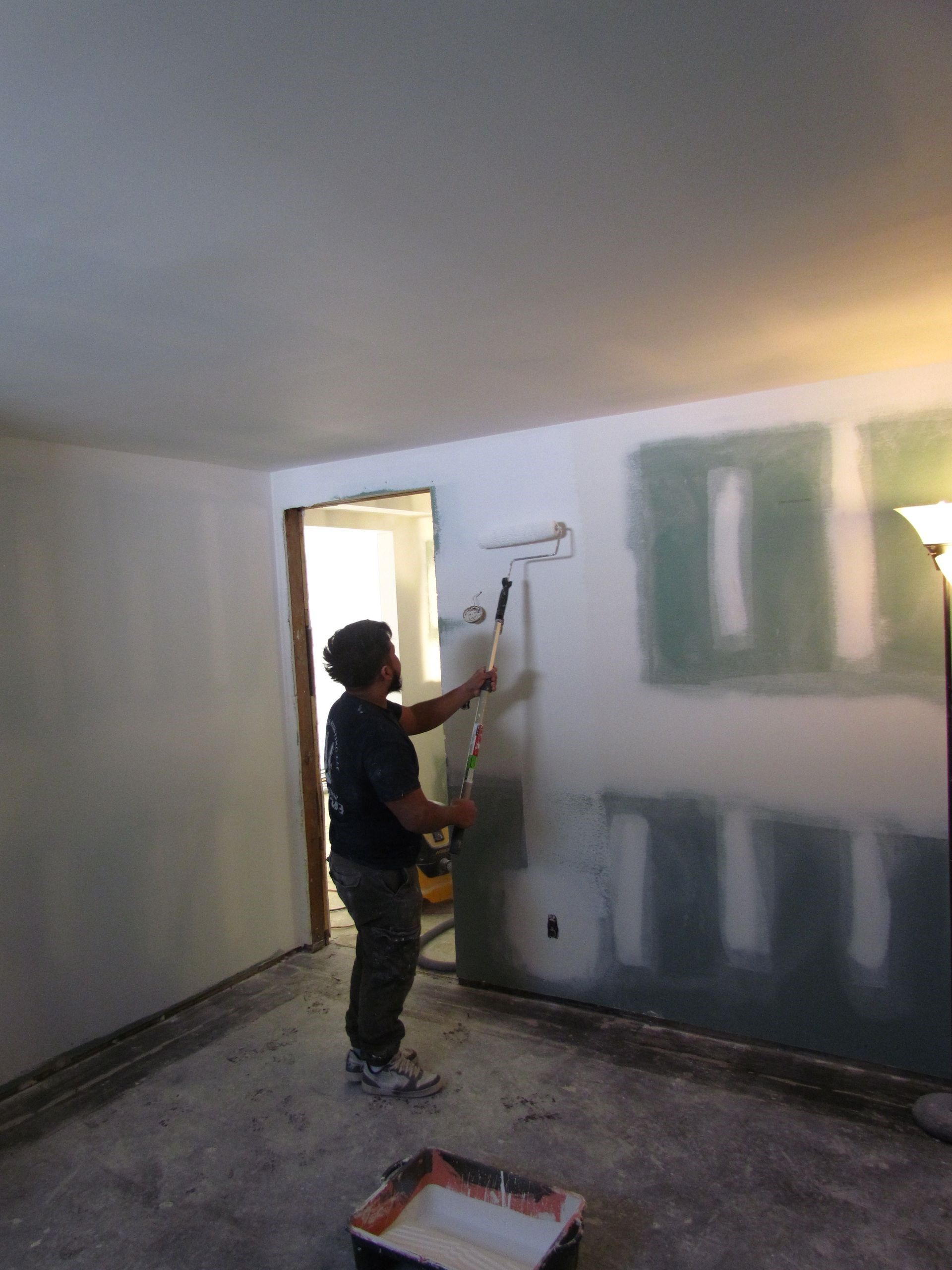 Person paints a wall with a roller; interior room with unfinished walls.